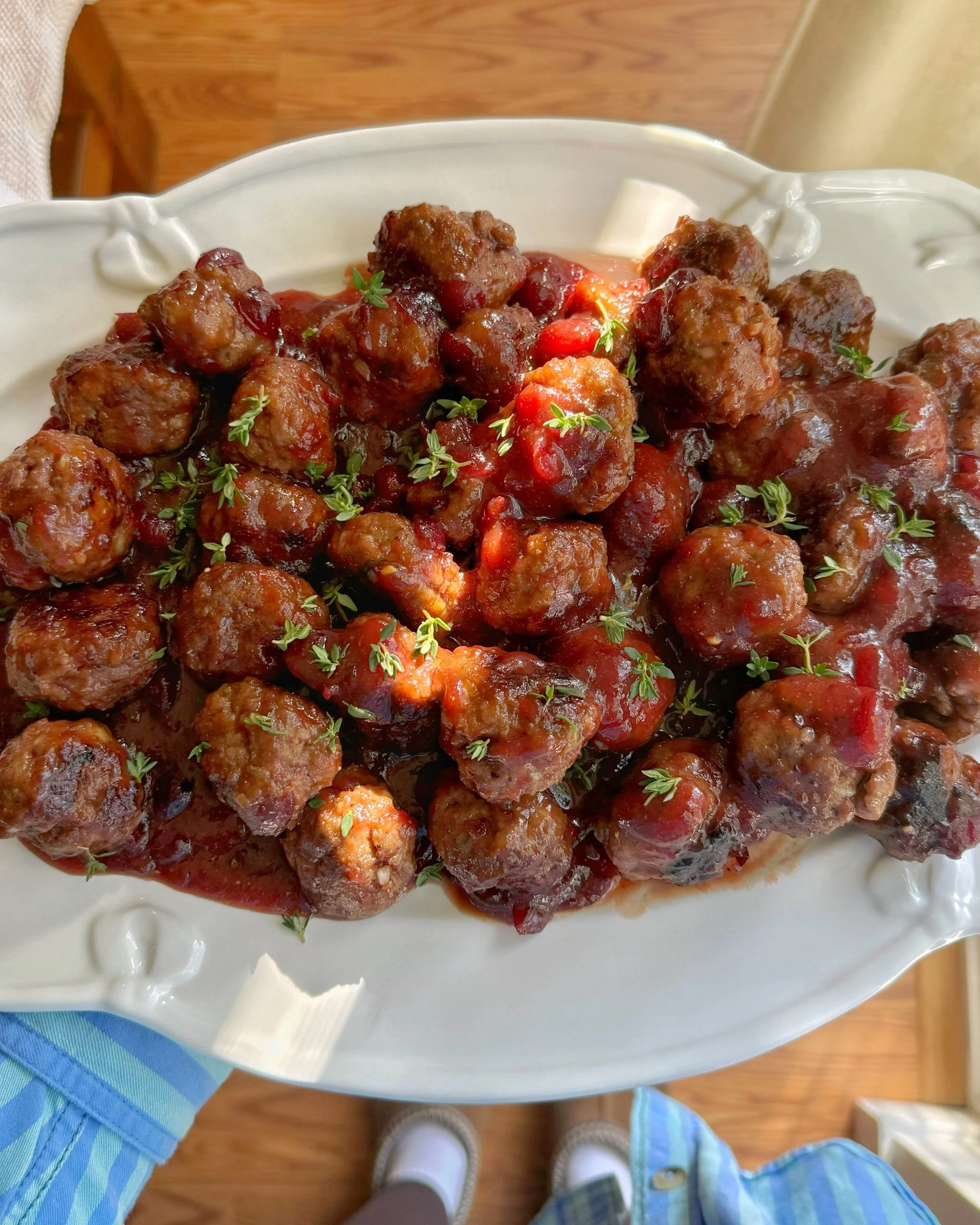 A platter of cranberry meatballs with sauce and  fresh thyme on top. 