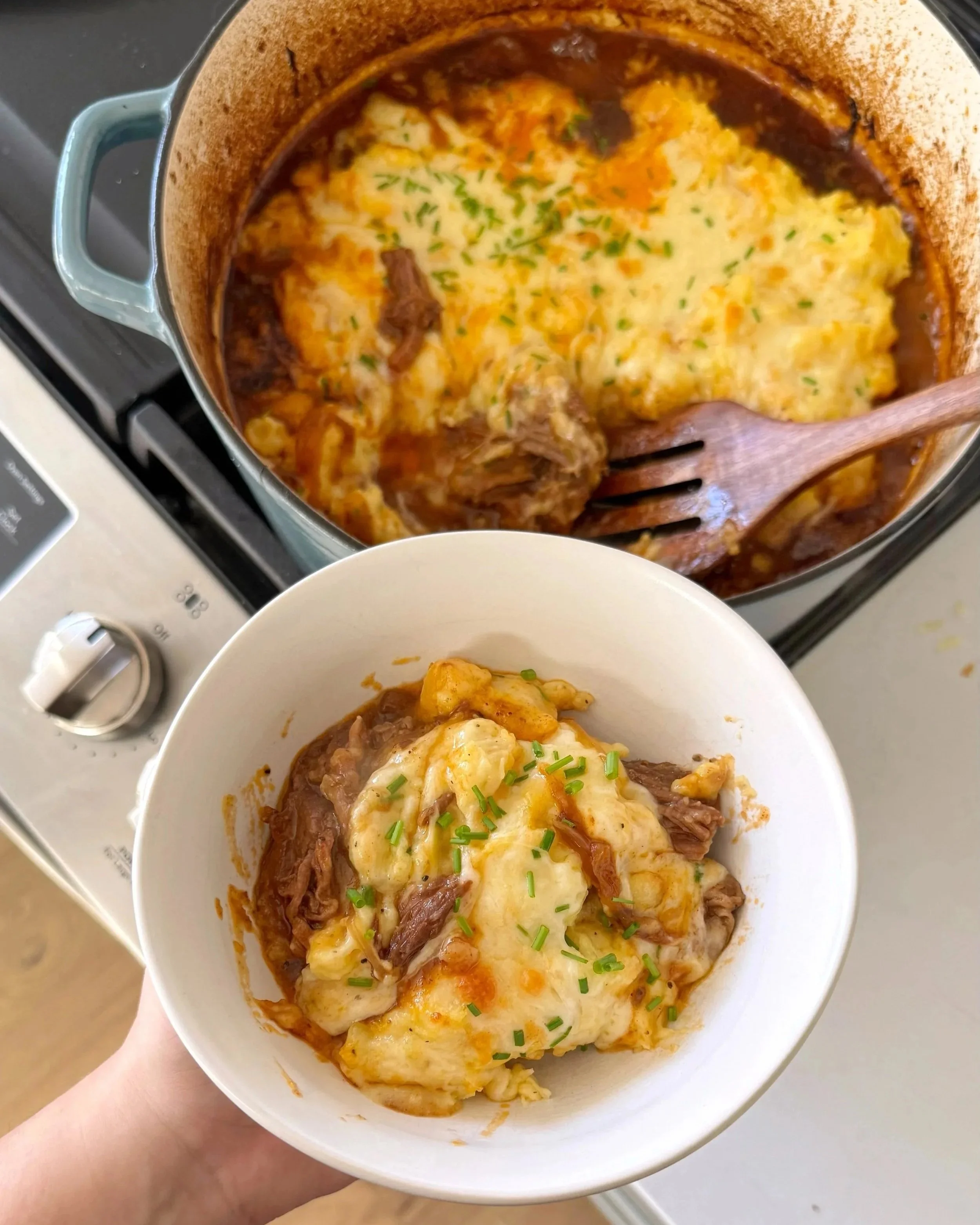 A pot of roast & mash potatoes with chives sprinkled on top with a bowl of the same thing being held up to the camera.