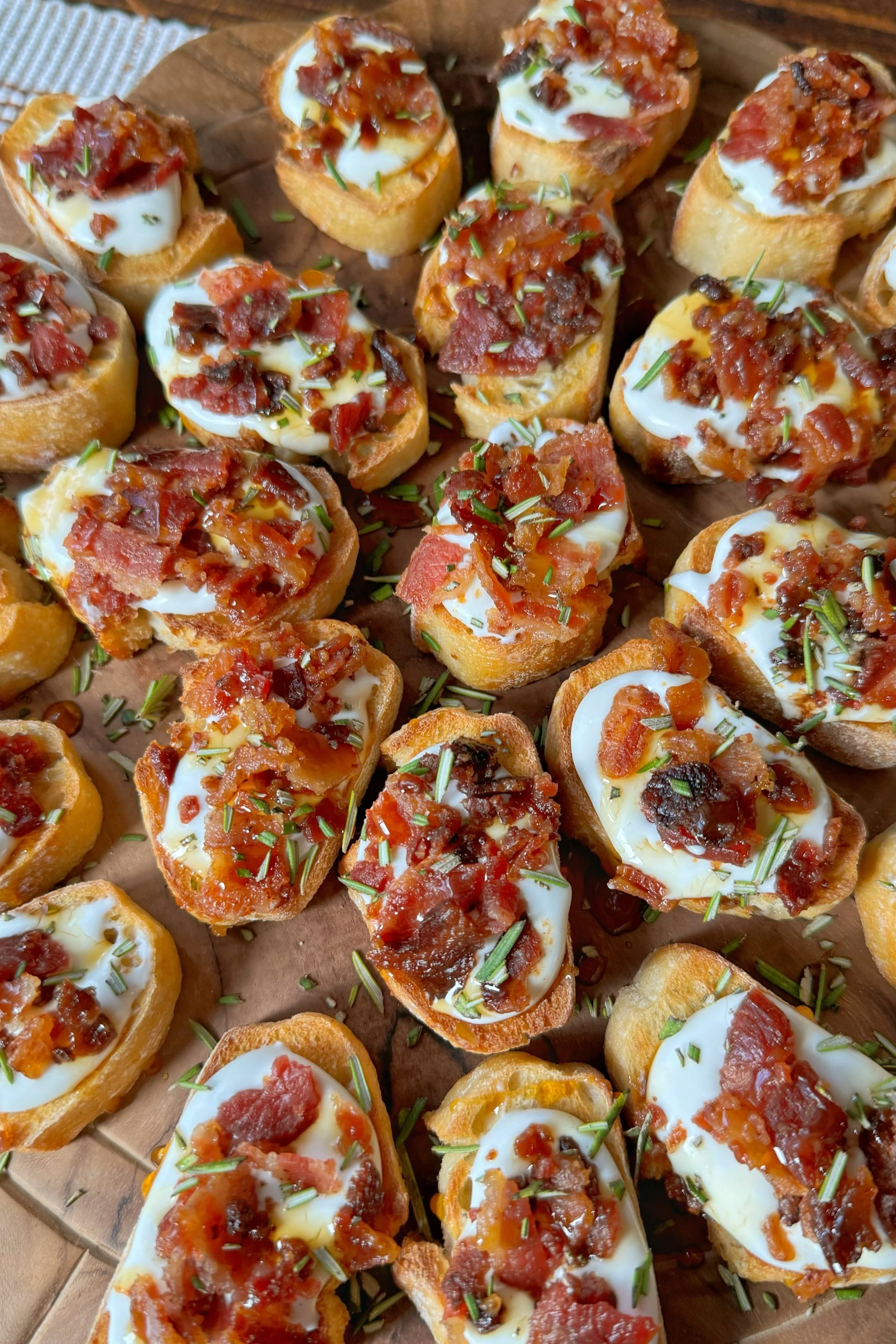 A cutting board full of little crostini with whipped goat cheese, bacon and chopped rosemary on top. Plus a drizzle of honey on each one.