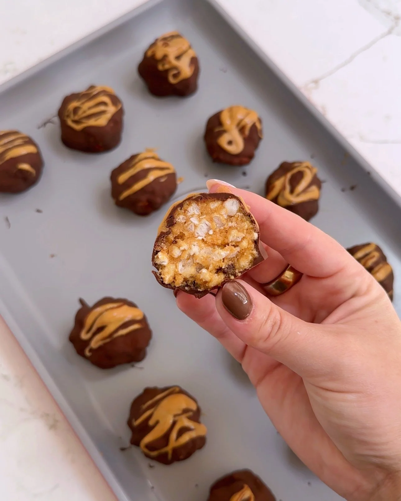 A sheet pan with chocolate bites with a drizzle of peanut butter on top. One bite is being held up to the camera to show the rice cake crunch inside the bite.