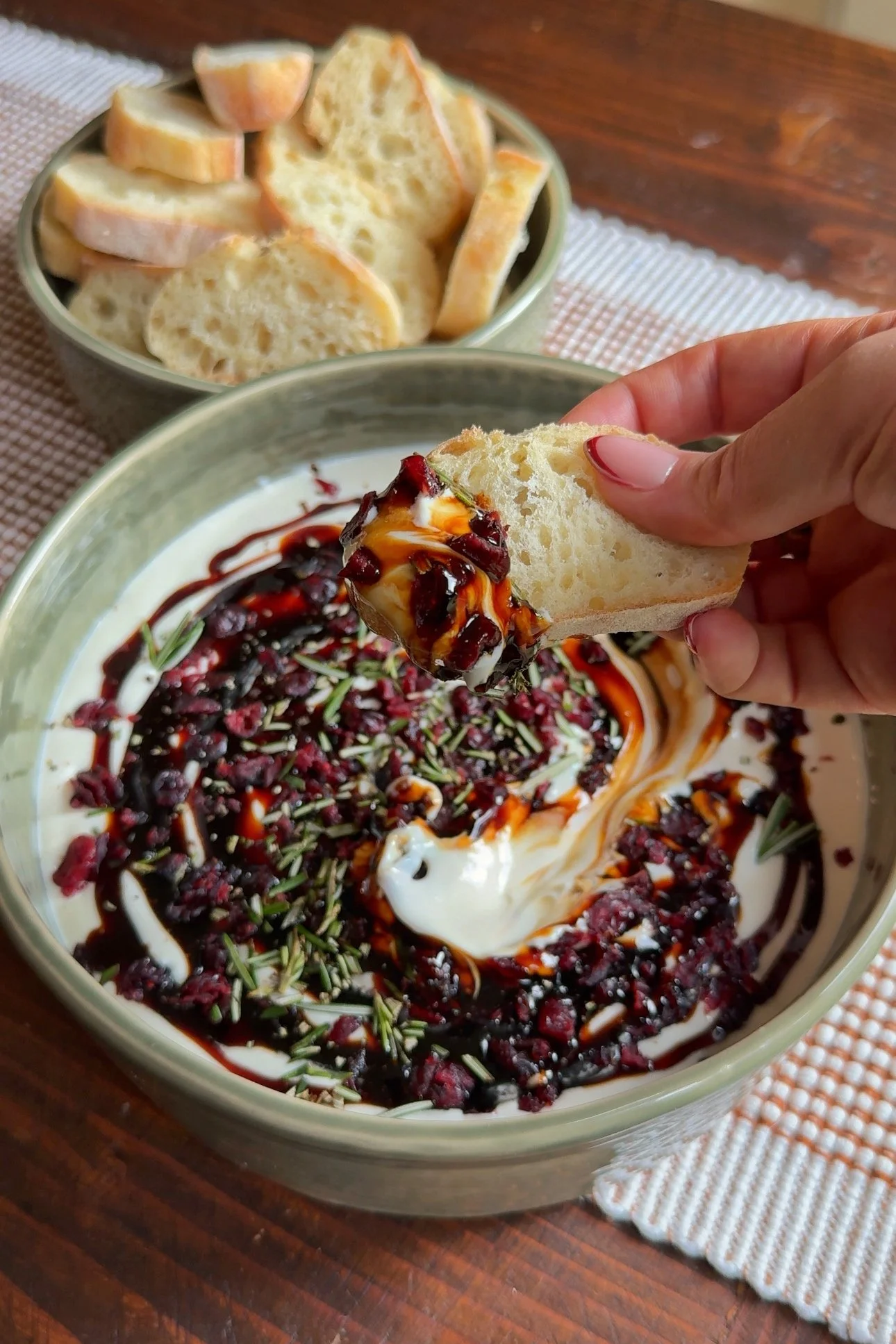 A bowl of whipped ricotta dip with balsamic, cranberries and fresh rosemary on top served with a side of bread. 