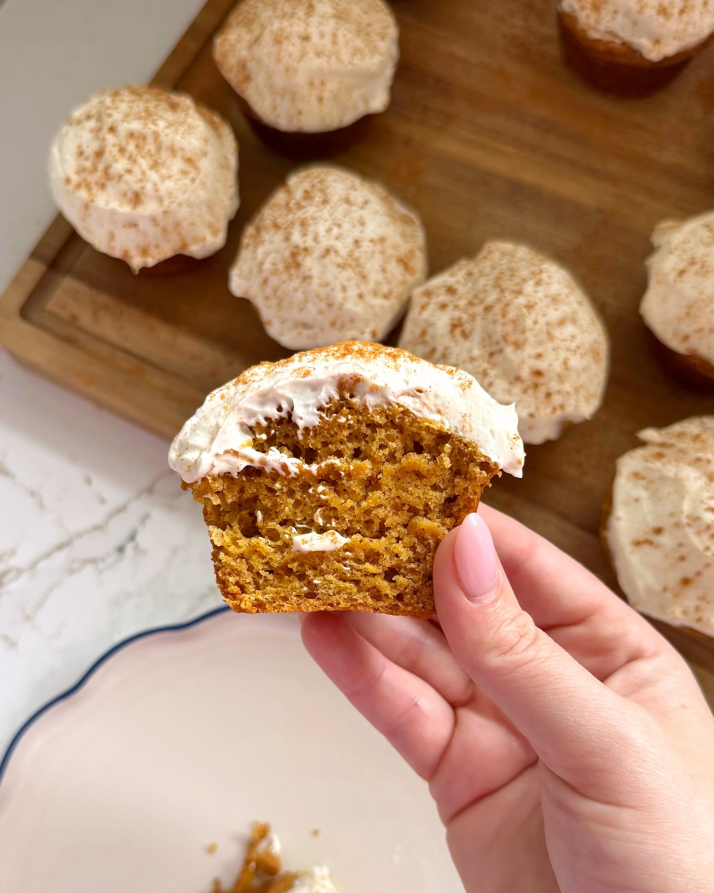 Pumpkin muffins with frosting sitting on top of a cutting board. One muffin is being held up to the camera. 
