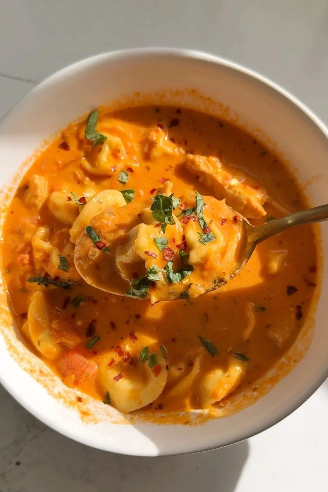 A close up shot of a spoonful of soup with crushed tomatoes, tortellini noodles, chicken and spinach topped with red pepper flakes and fresh basil.