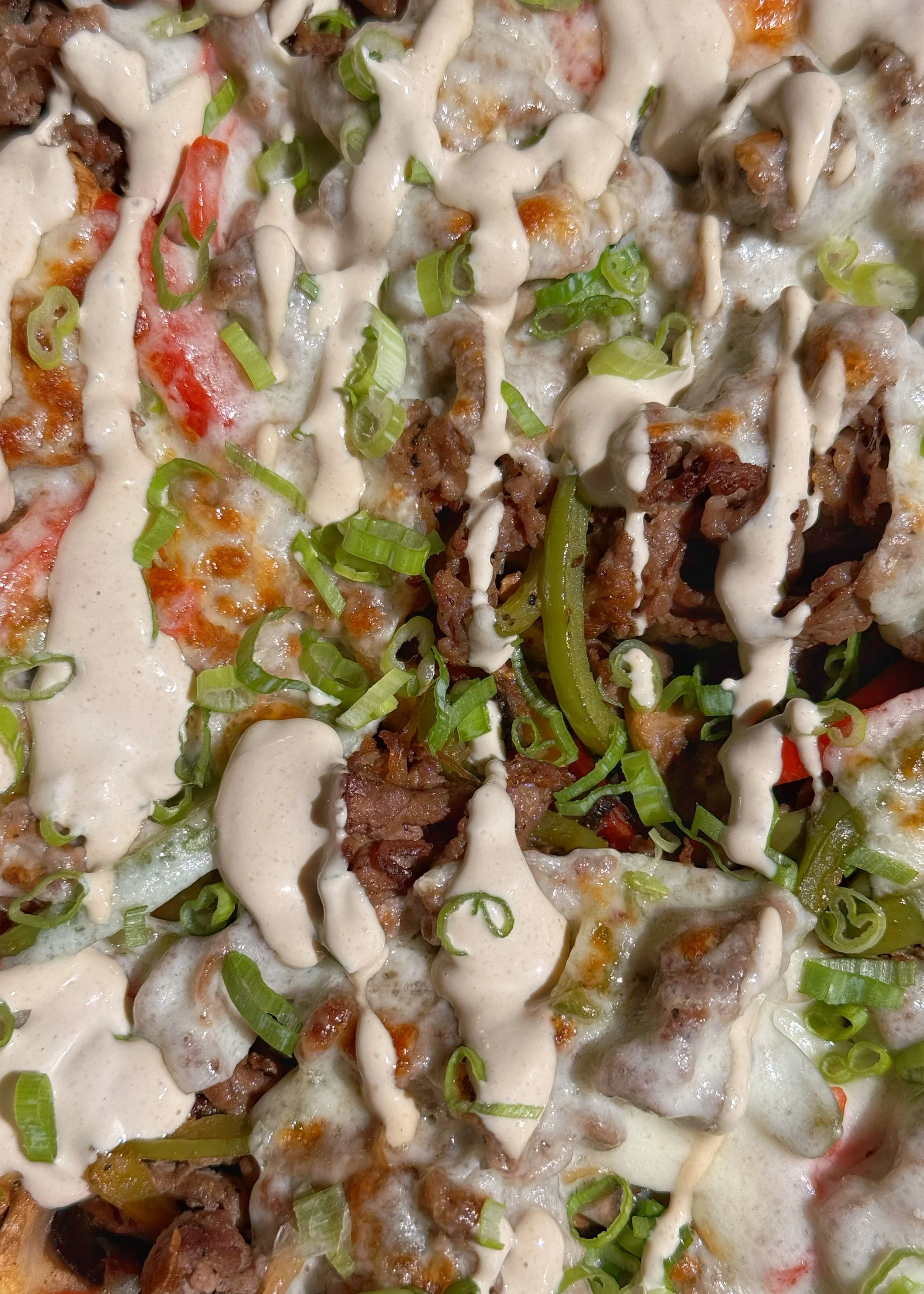 A close up shot of cheesesteak, bell peppers & mushrooms with chopped green onions and a dressing drizzled on top.