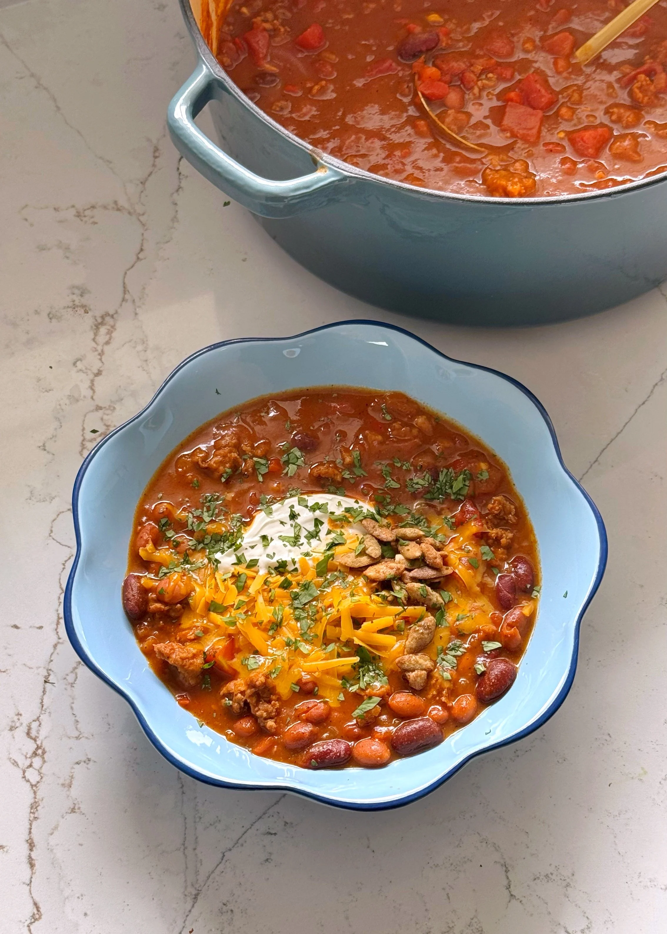 Sitting next to a big pot with pumpkin chili is a bowl of pumpkin chili topped with sour cream, cheese, pumpkin seeds and chopped cilantro.