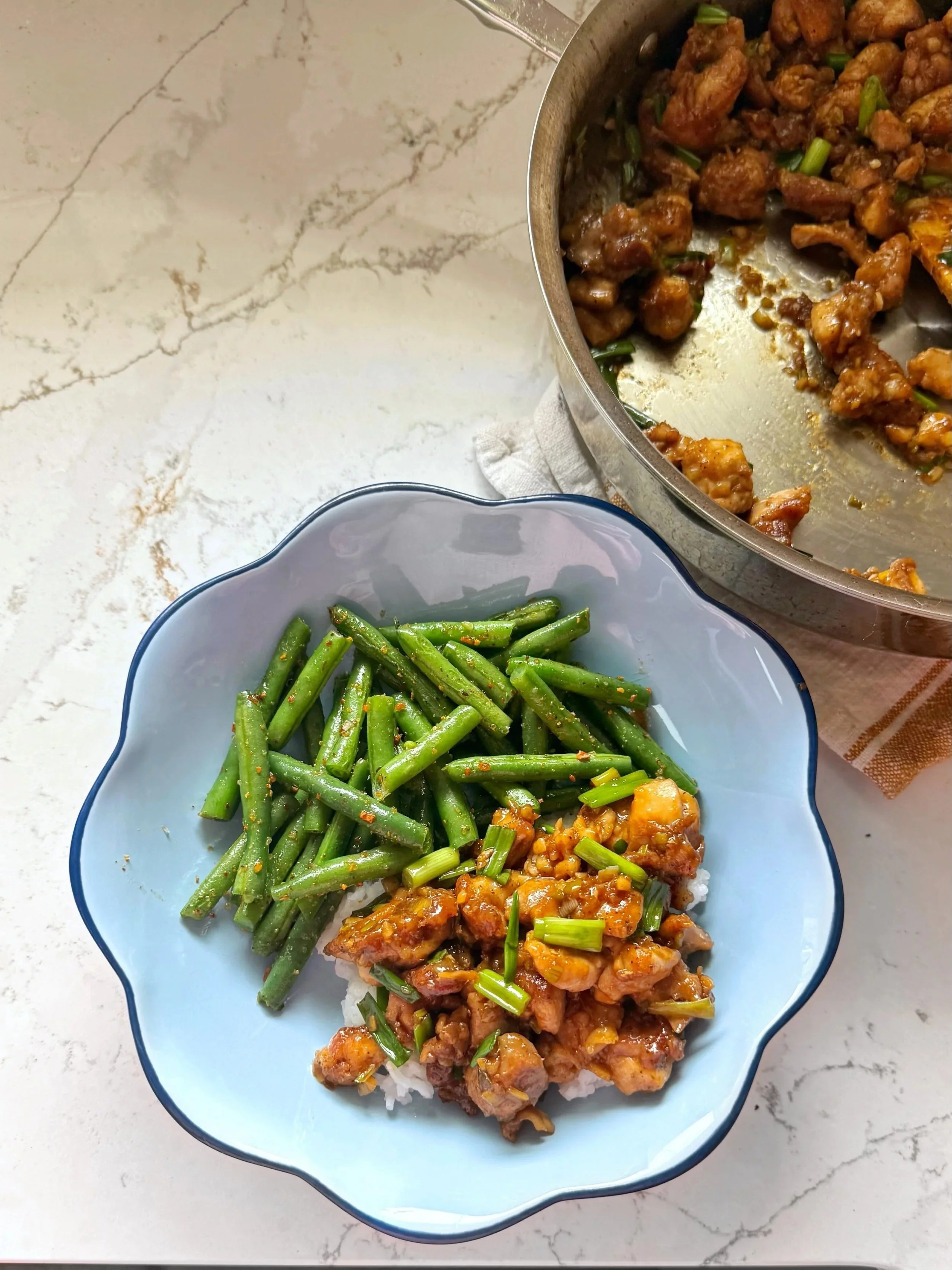 A bowl of scallion chicken with a side of green beans sitting next to a pan of chicken, scallions, & garlic with a glaze sauce. 