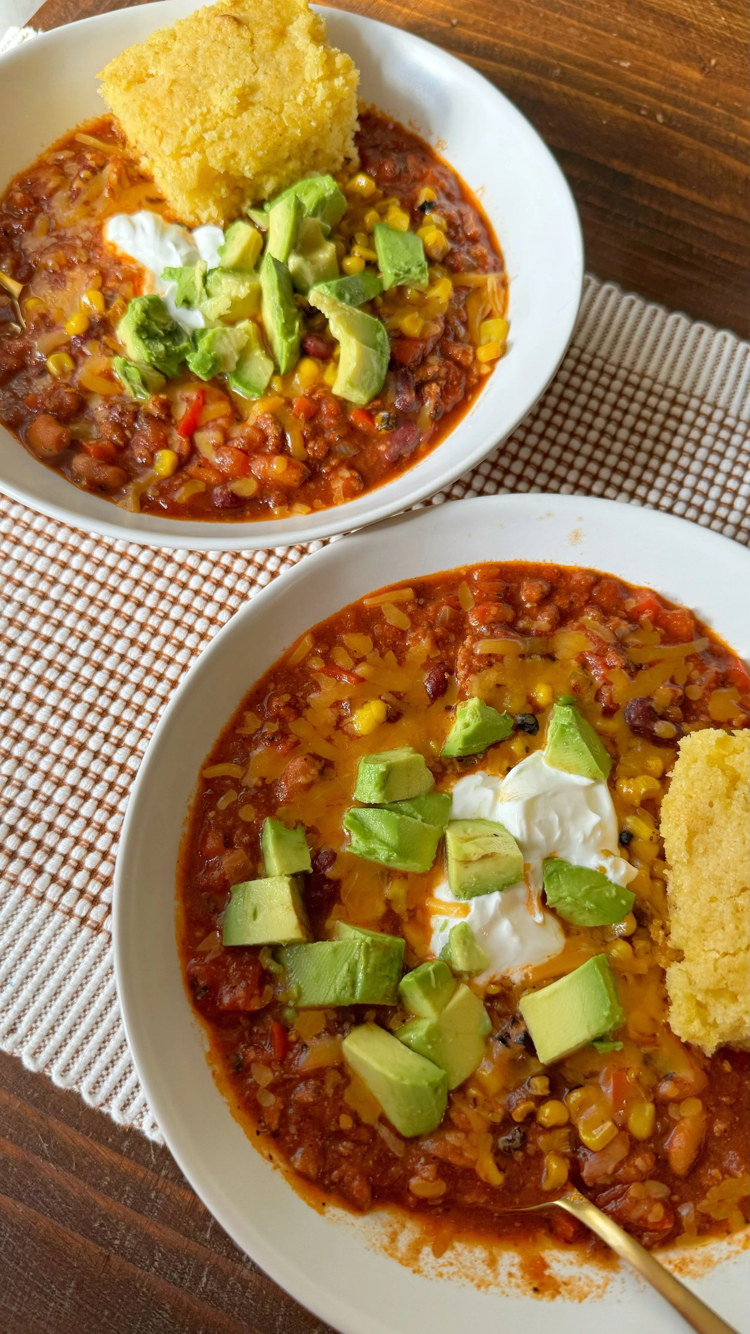 Two bowls of chili with avocado and sour cream on top with a side of cornbread.