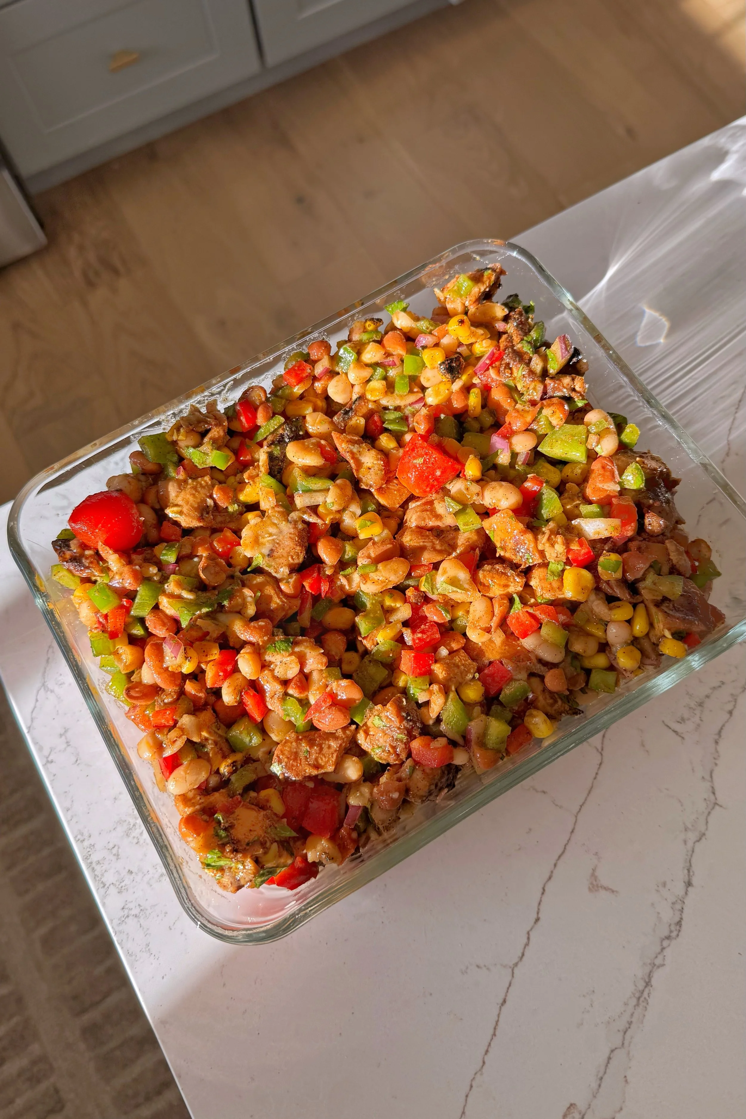 A glass pyrex dish with the bean salad that includes chicken chunks, tomatoes, corn, red onion, white beans, and green peppers.