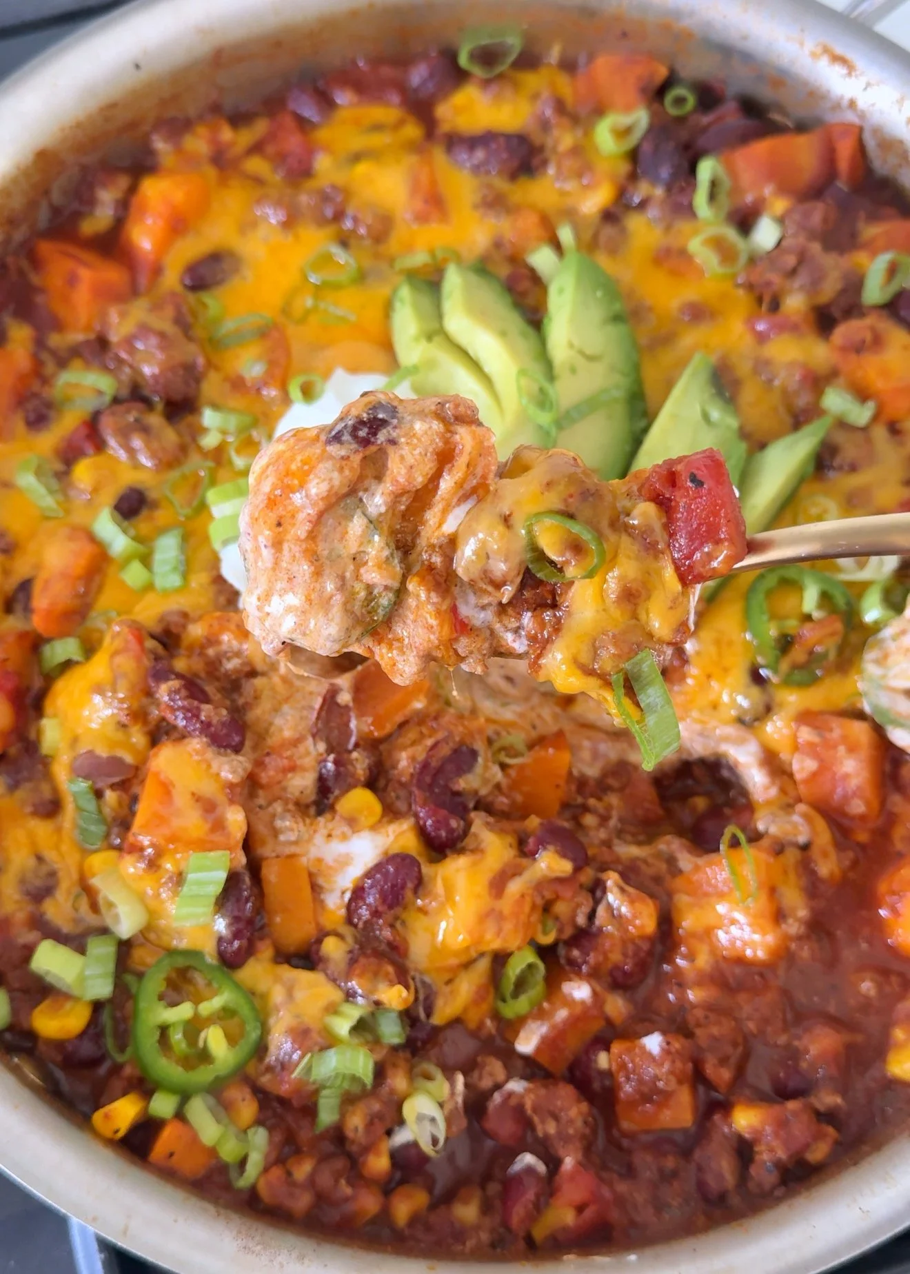 A close up shot of a pan with sweet potato chili in it topped with fresh avocado, sour cream, chopped onions and Jalapeño slices. 