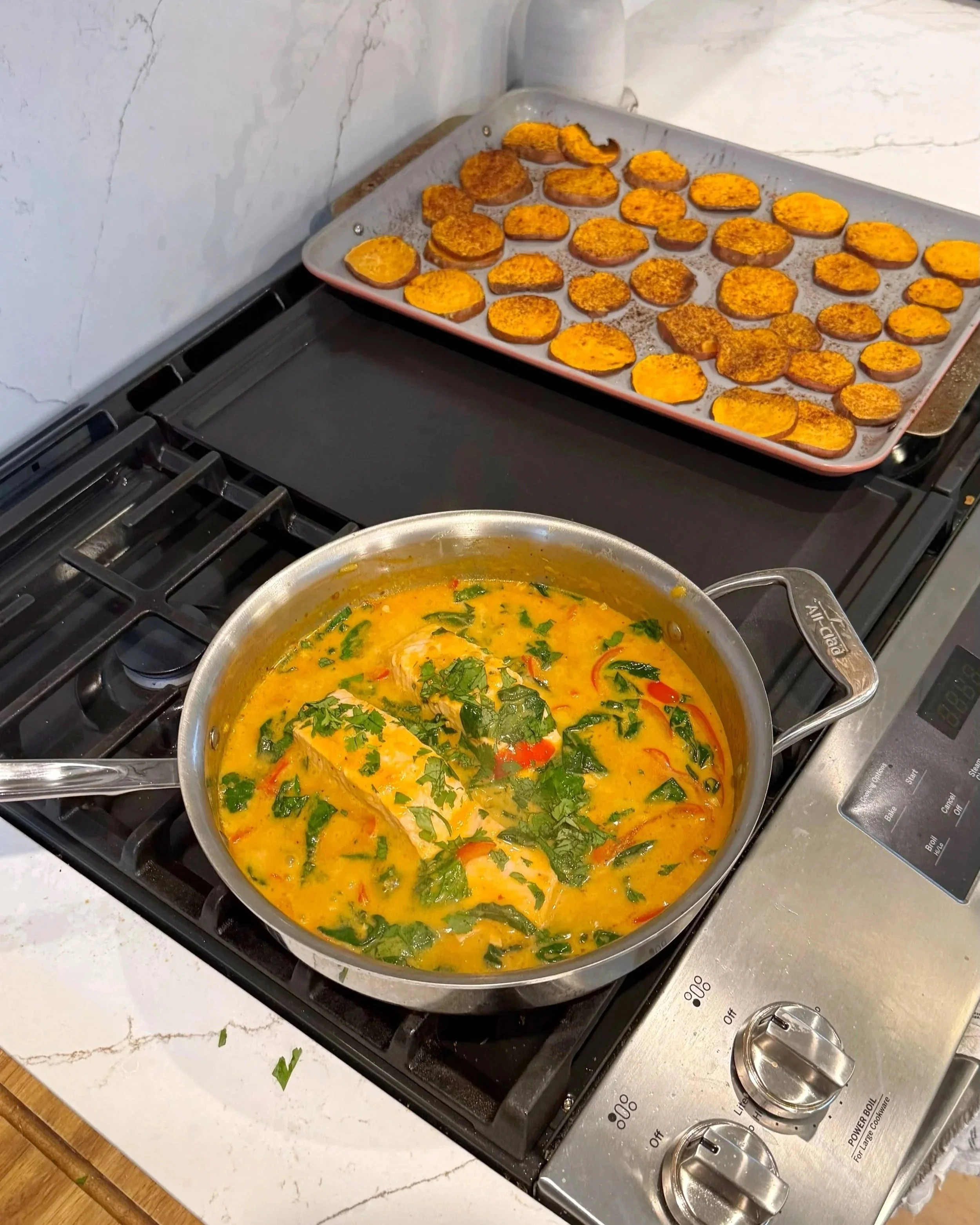 A pan of coconut curry salmon with chopped red pepper and cilantro with a side of sweet potato circles on a baking sheet. 