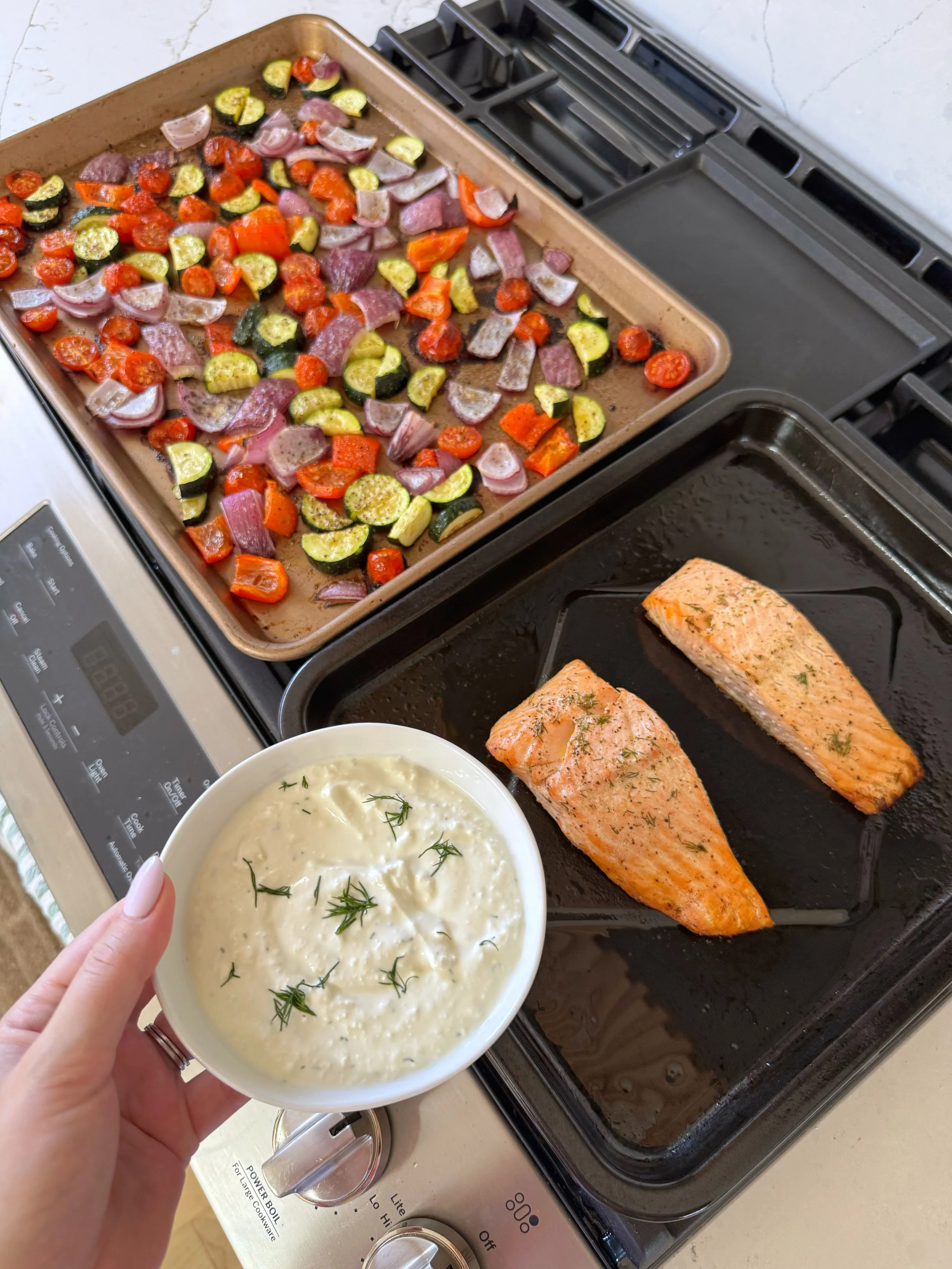 A pan of mixed roasted veggies next to a pan with 2 pieces of salmon and a dish with sauce and fresh dill on top.