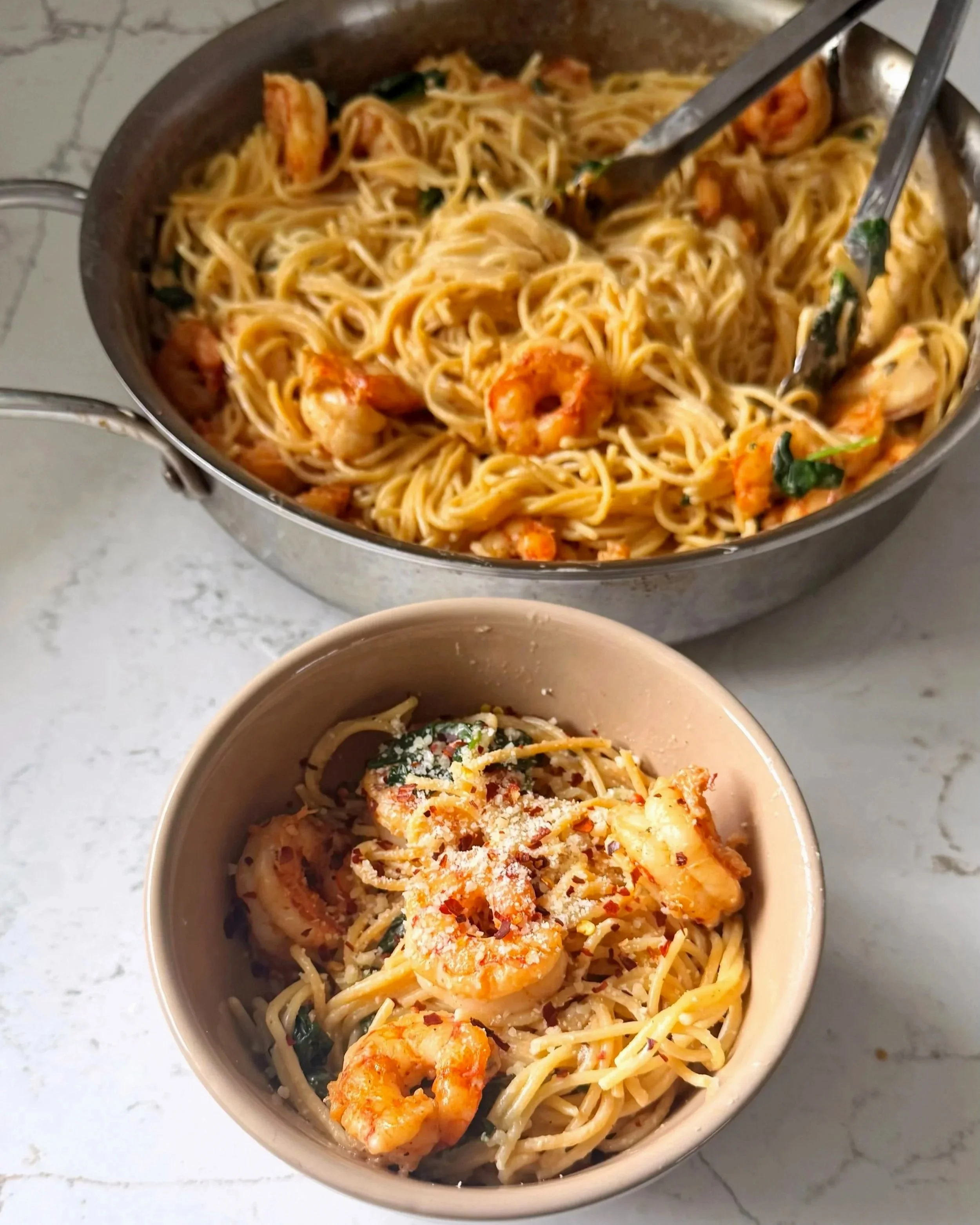 A bowl with pasta, shrimp, and spinach with a creamy sauce topped with red pepper flakes and grated cheese. It's sitting next to a pan with the same ingredients.