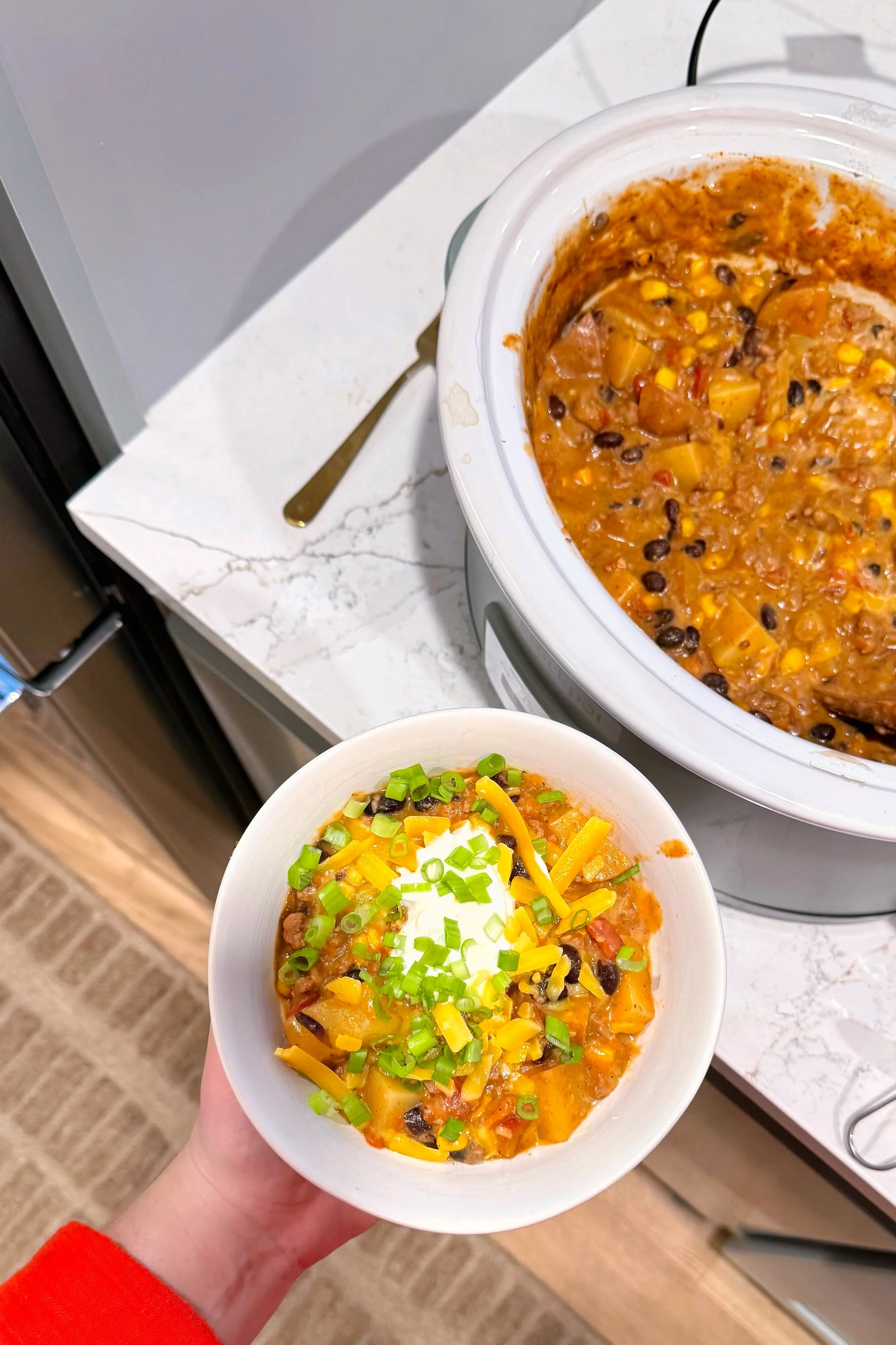 A bowl of cowboy casserole topped with sour cream, shredded cheddar cheese, and chopped green onions. It is sitting next to the crockpot with cowboy casserole in it.