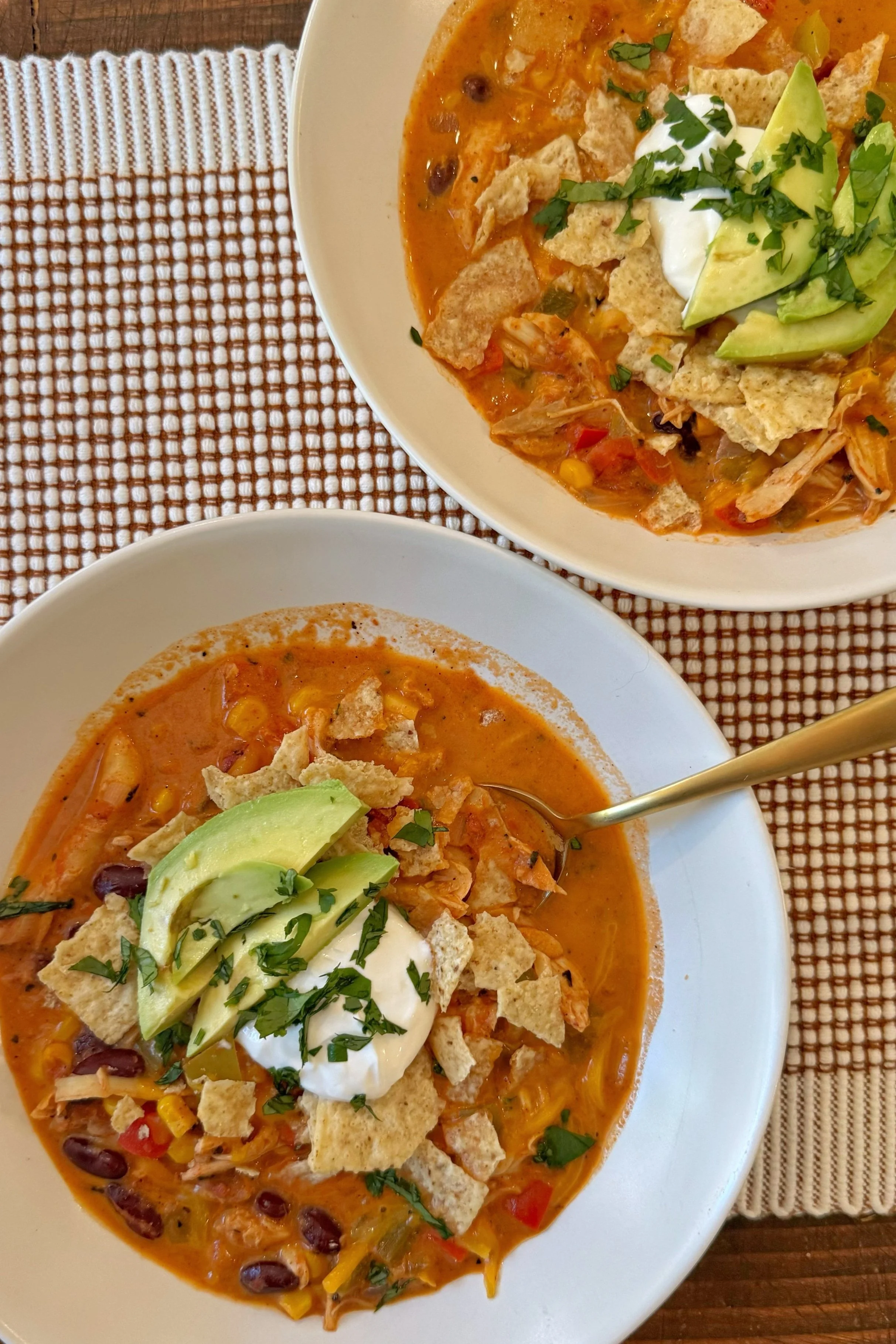 Two bowls of soup with taco ingredients in it topped with avocado, sour cream, corn chips and cilantro.