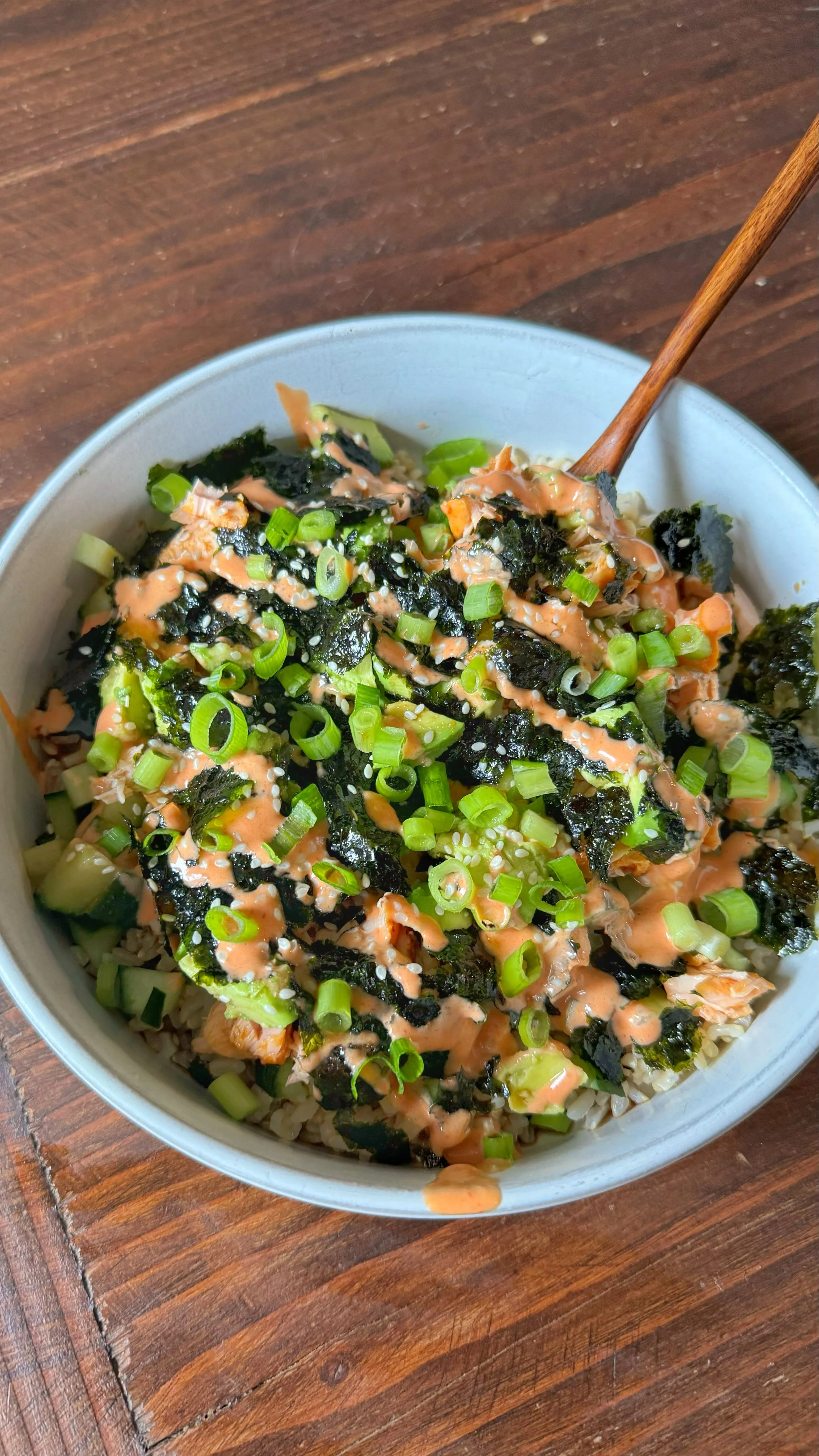 A bowl of salmon sushi with cucumbers, avocados, seaweed & brown rice topped with chopped green onions and sesame seeds.