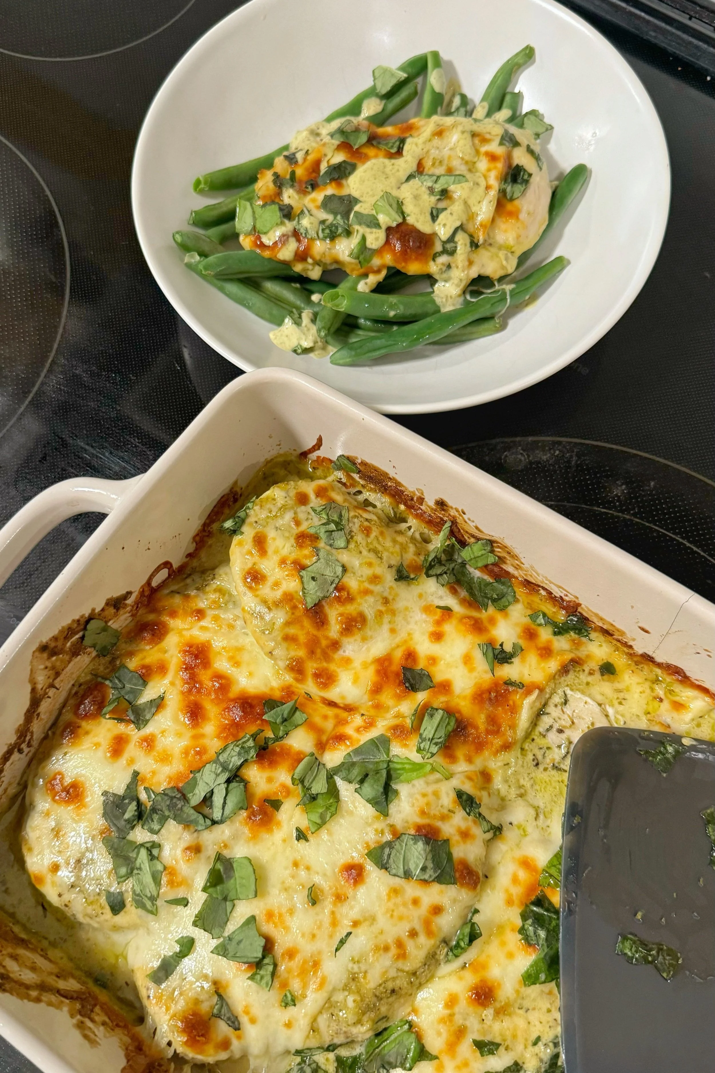 A baked pesto aioli chicken plated on top of cooked green beans sitting next to the baking dish with the chicken bake.