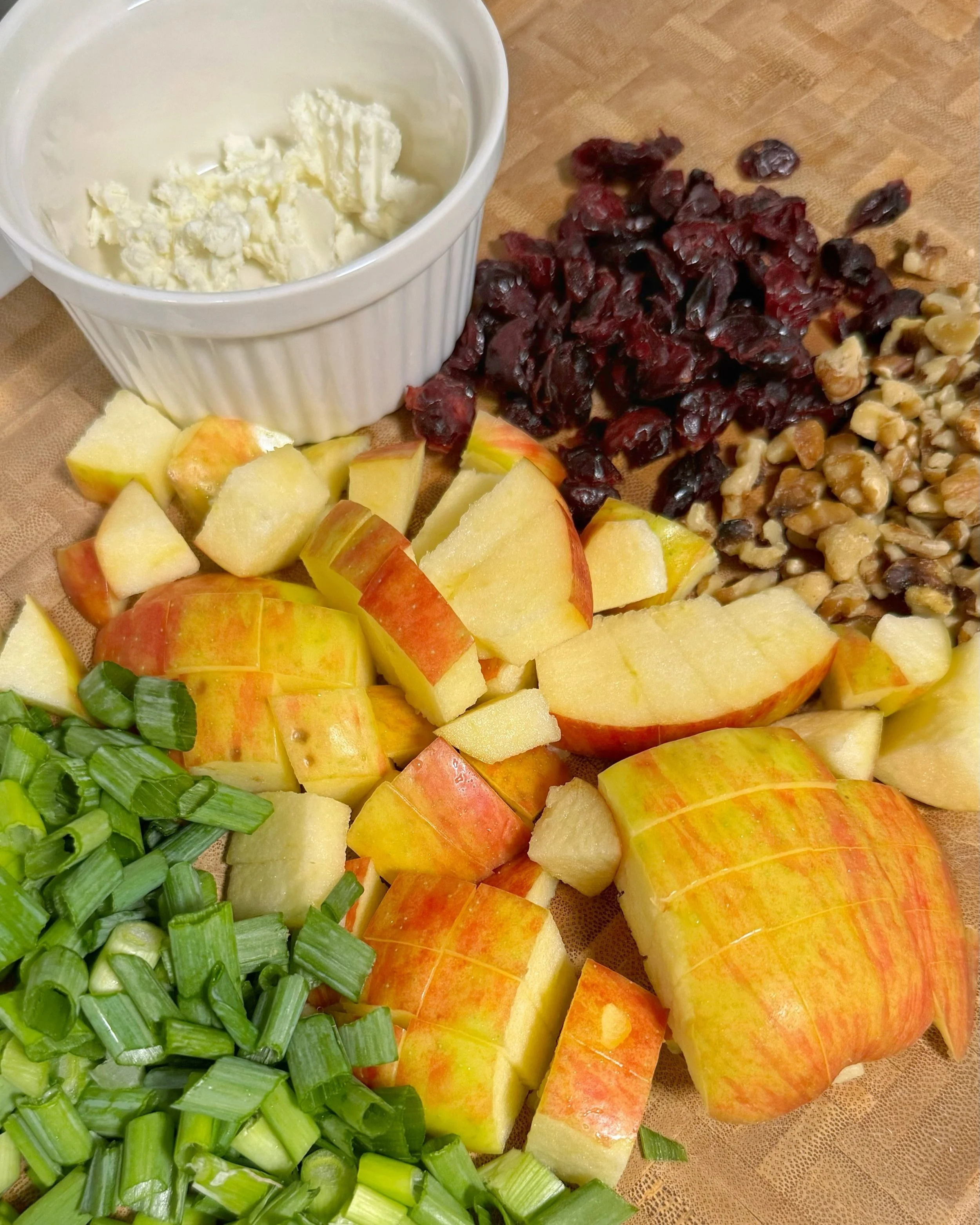 Apple chunks, chopped green onions, walnuts, dried cranberries on top of a cutting board with a side of goat cheese.