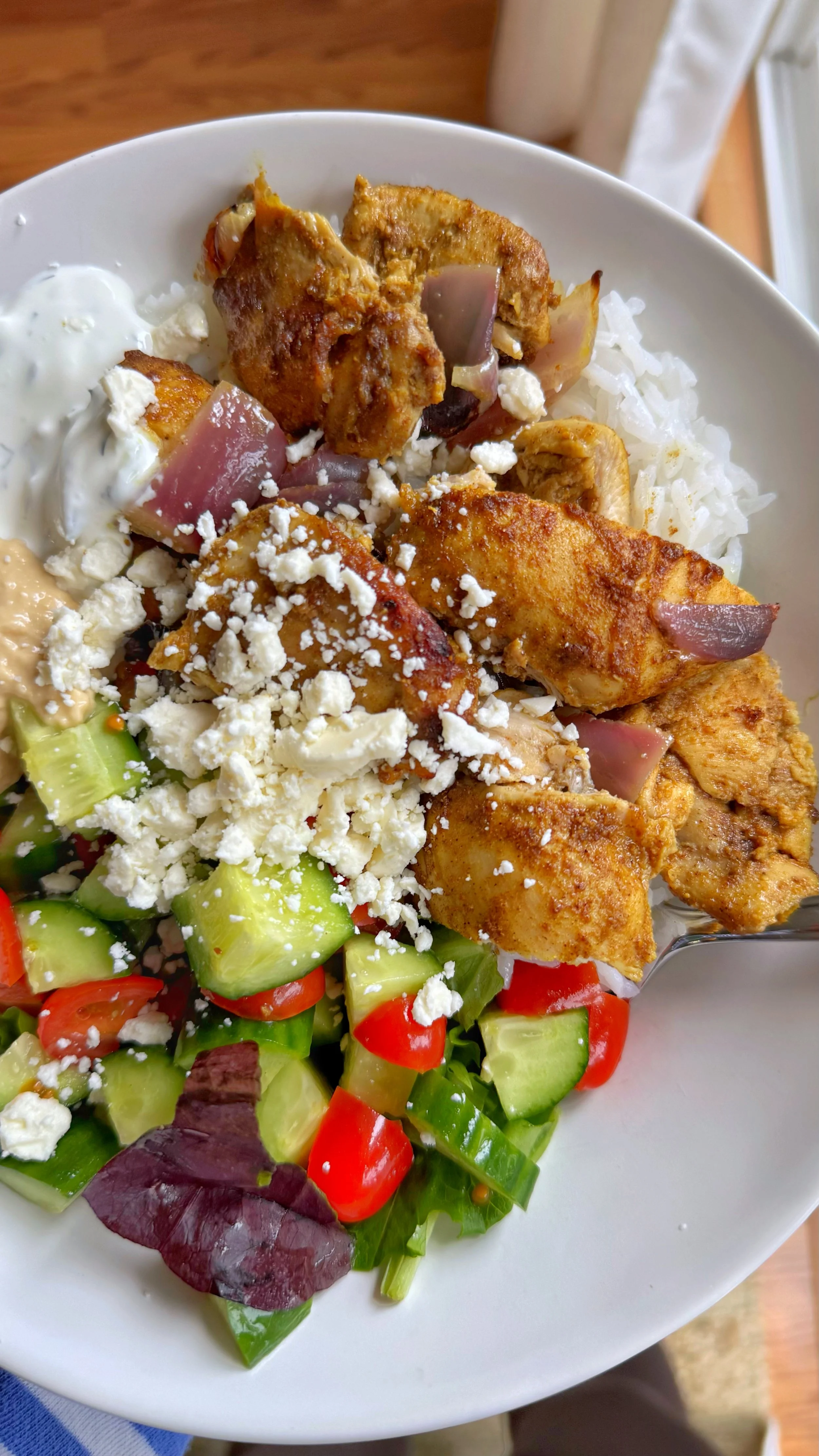 A close up picture of a bowl of chicken shawarma with cucumber, tomato, hummus, and tzatziki on a bed of rice with feta cheese on top.