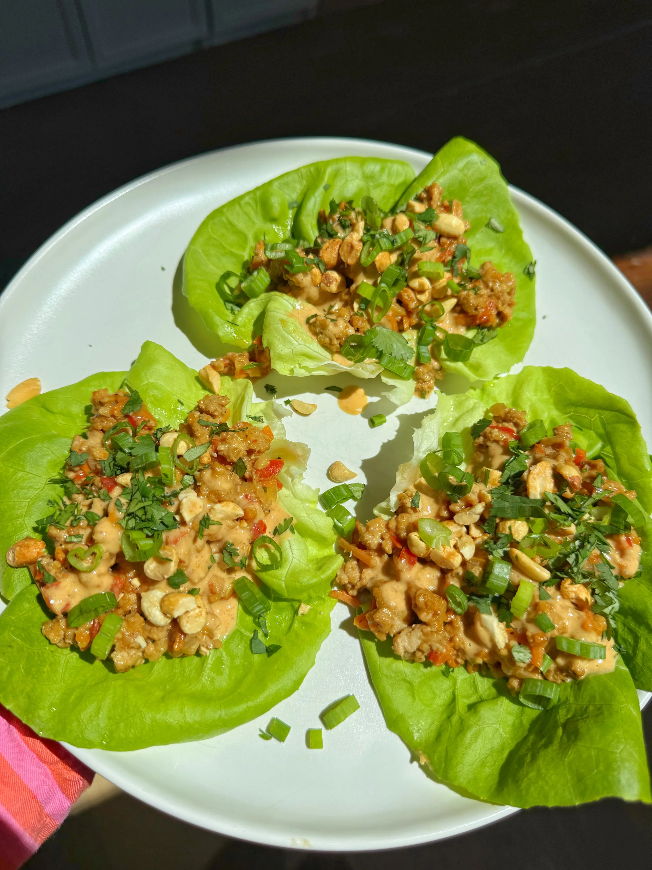 A plate of 3 lettuce wraps with Thai peanut chicken in it topped with chopped green onion and cilantro. 