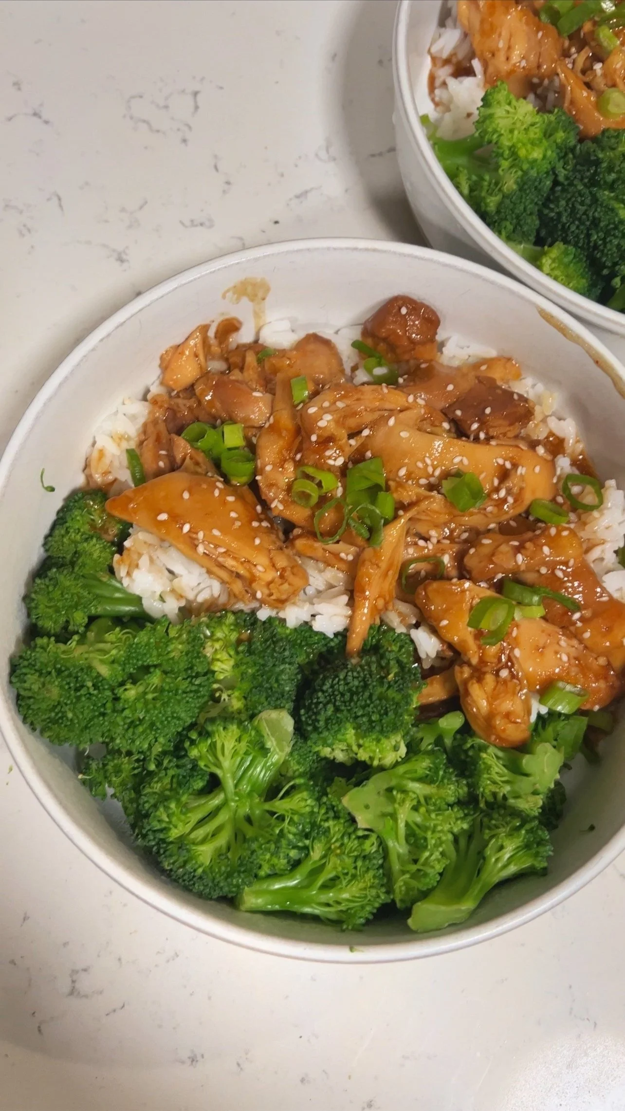 A bowl of honey chicken with garlic & broccoli on a bed of rice topped with green onions and sesame seeds.