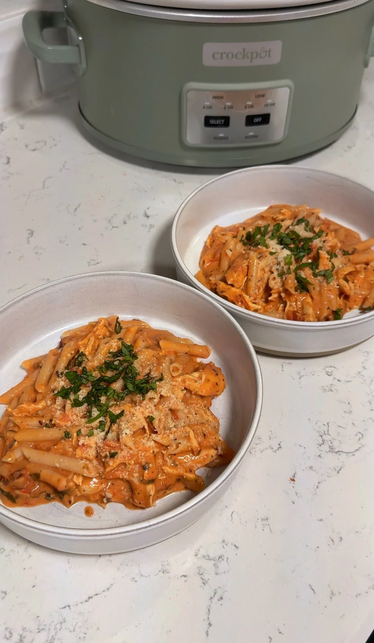 Two bowls of tomato chicken pasta with basil on top. Sitting next to a crockpot.