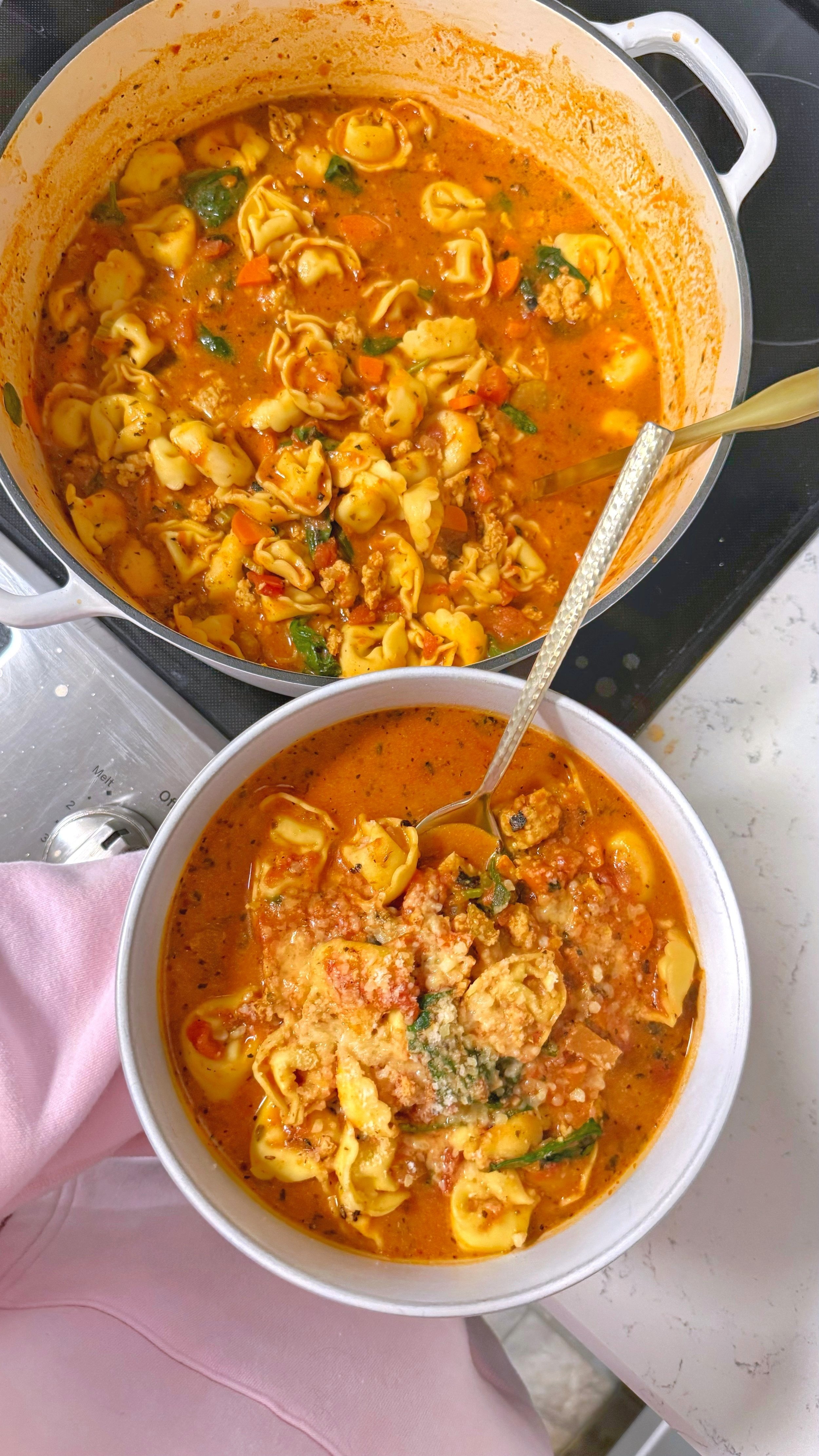 Sitting next to a pot of soup is a bowl of tortellini soup with spinach in a tomato based broth with grated parmesan on top.