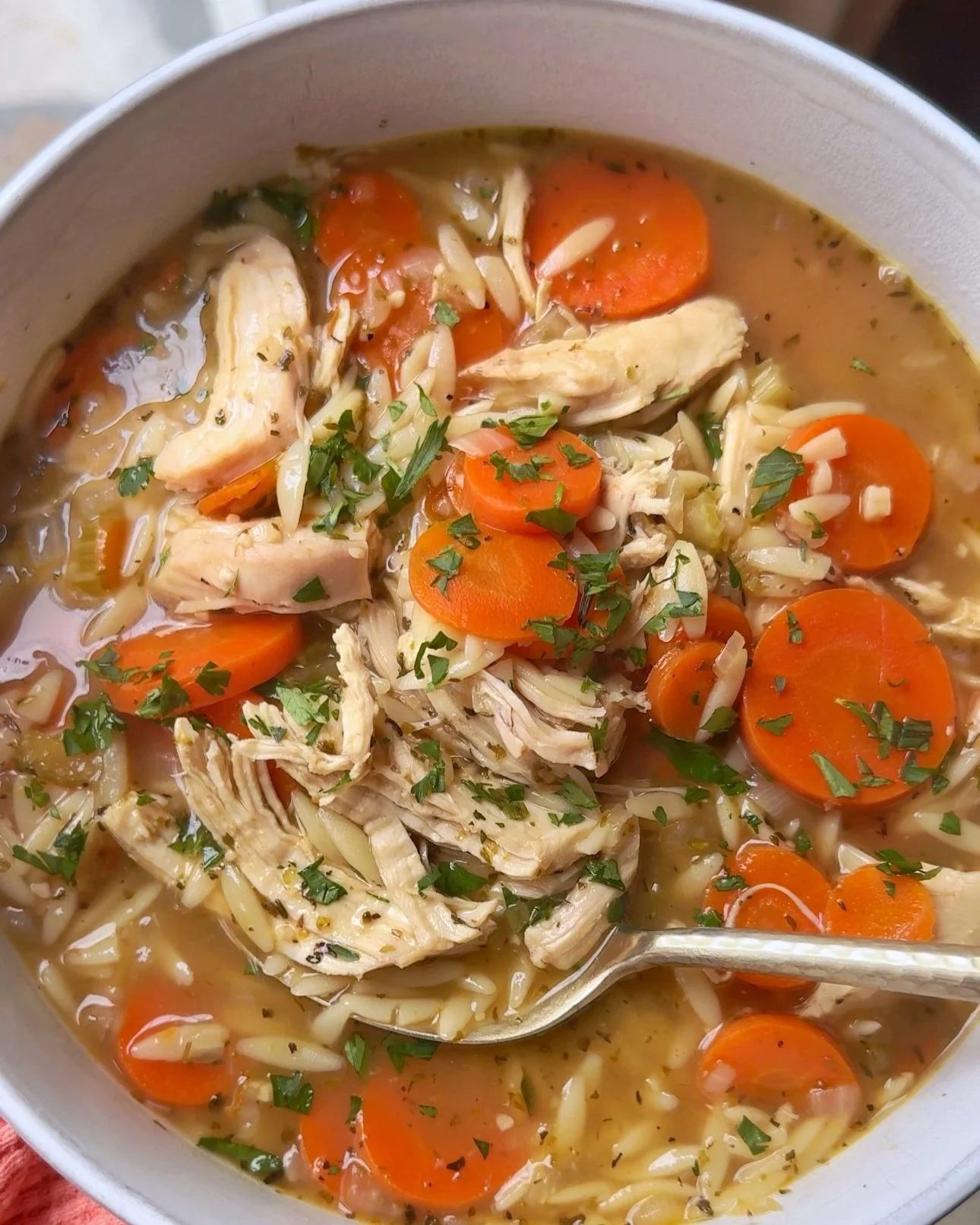 A close up shot of a bowl of chicken and orzo soup with carrots & onions topped with fresh parsley.