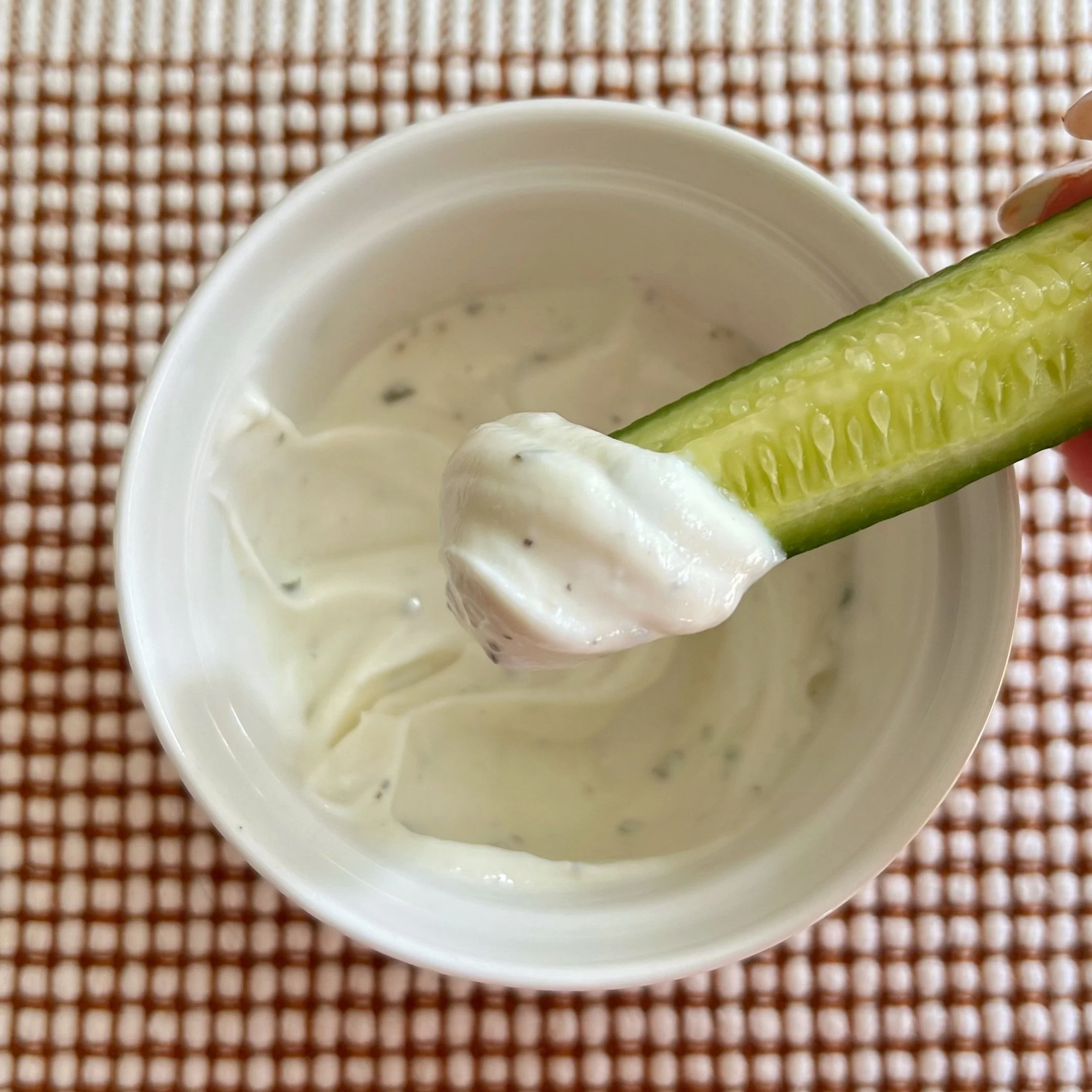 A small bowl of ranch dip with a cucumber dipped into it.