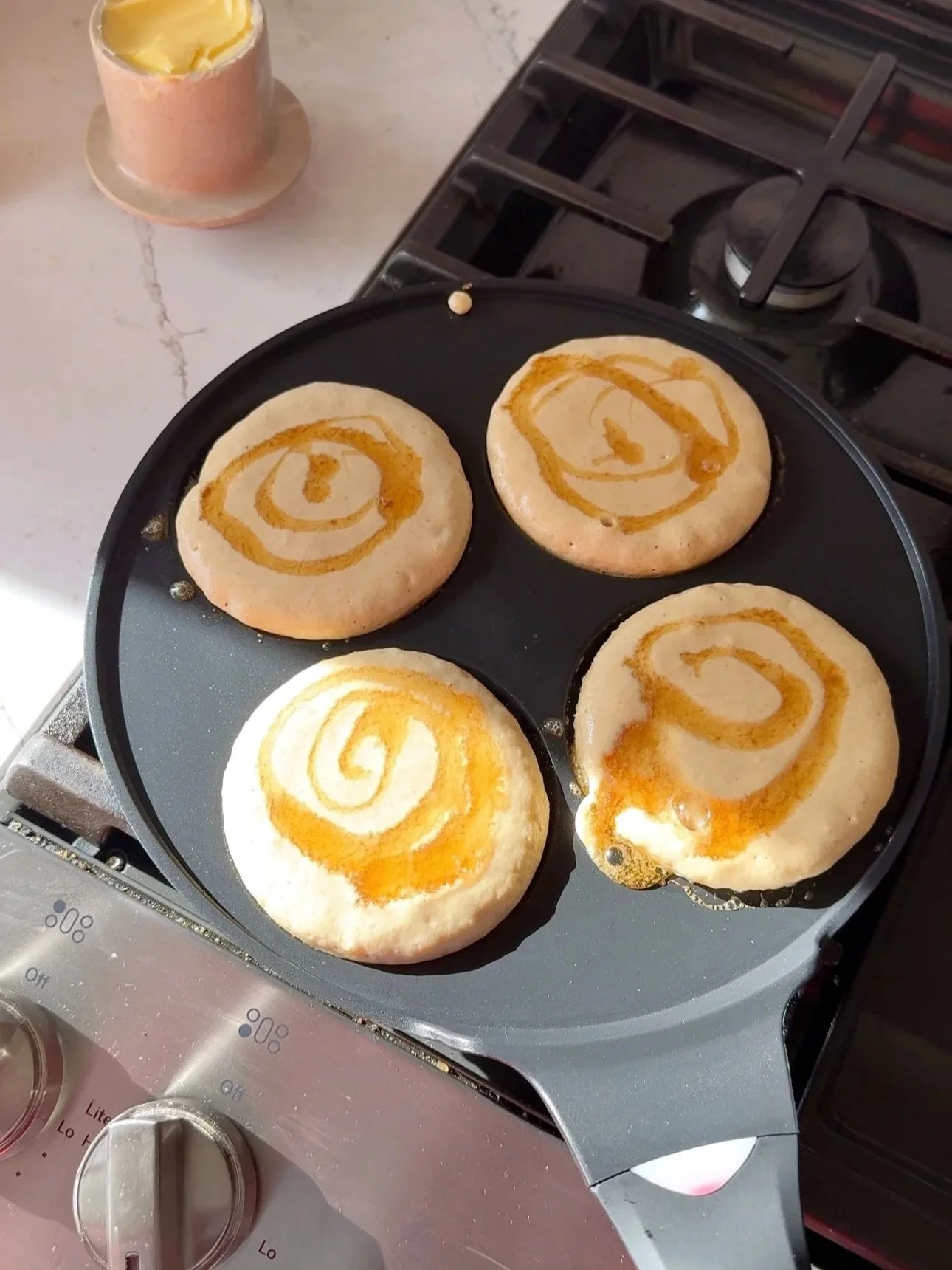 A pan with pancakes cooking. Pancakes have a drizzle of maple syrup on them. 