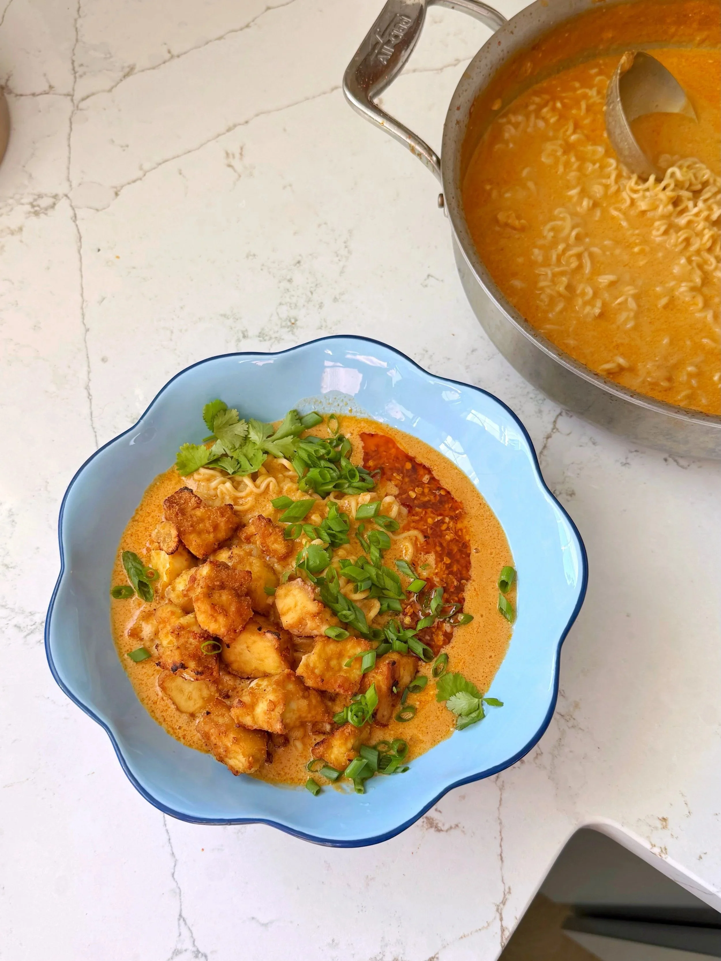 A bowl of Tofu and ramen in a creamy sauce topped with fresh chopped green onion and cilantro. Sitting next to a pan with creamy ramen in it.