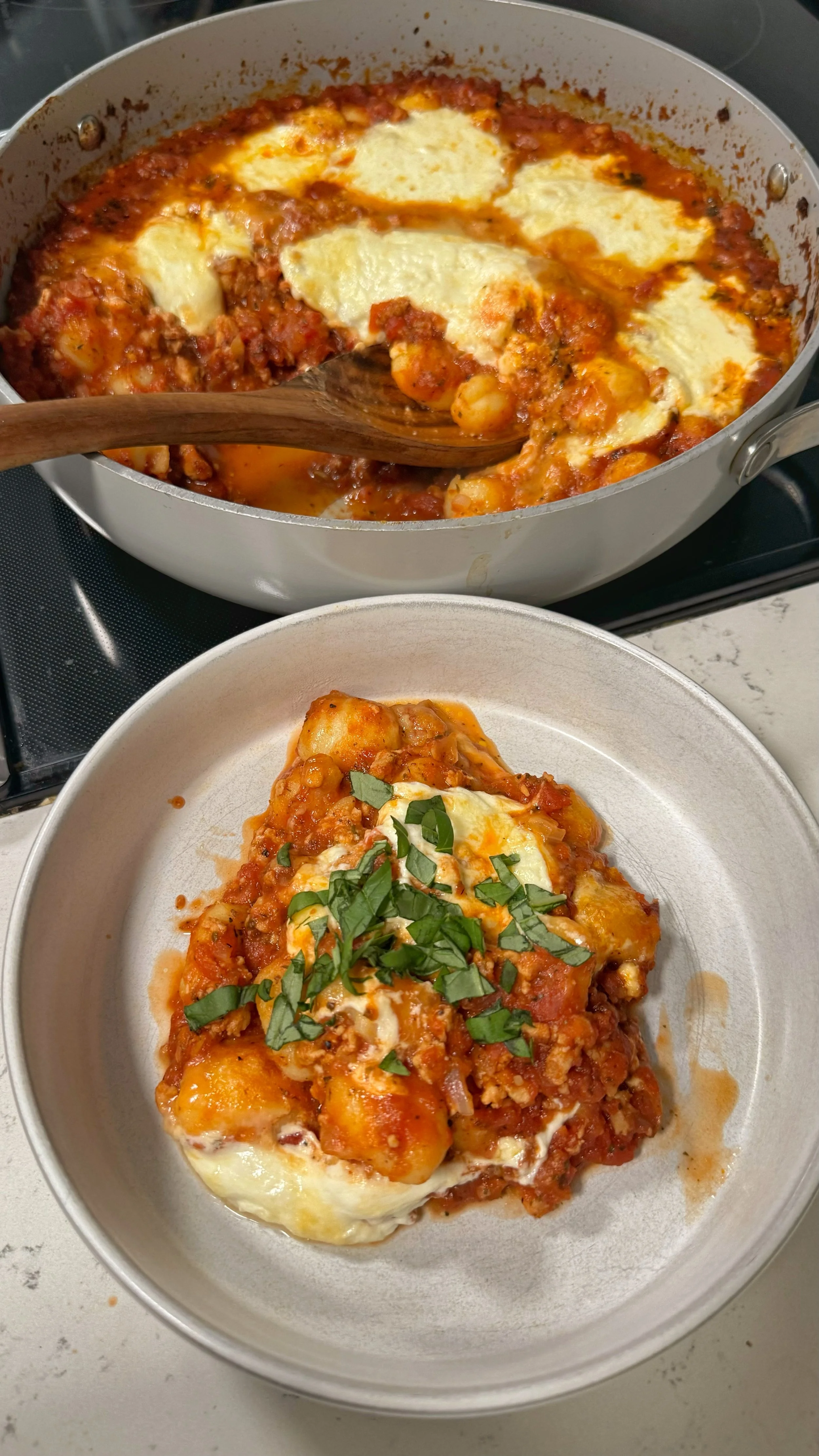 A pan with baked gnocchi in a tomato meat sauce & cheese. Sitting next to it is a serving of it in a bowl topped with fresh basil. 