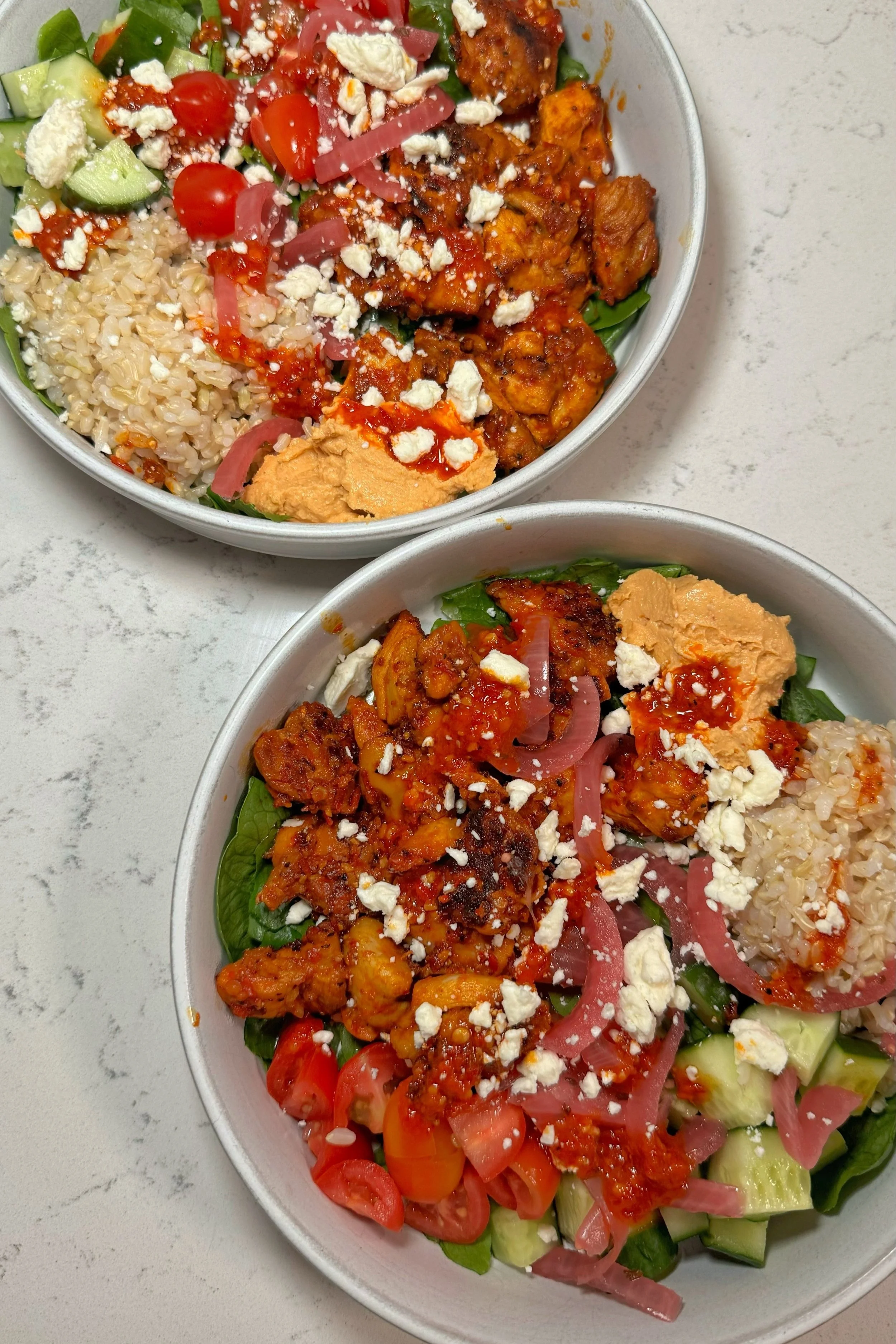 Two bowls of chicken, rice, pickled onions, cucumbers and feta cheese on top of a bed of mixed greens.