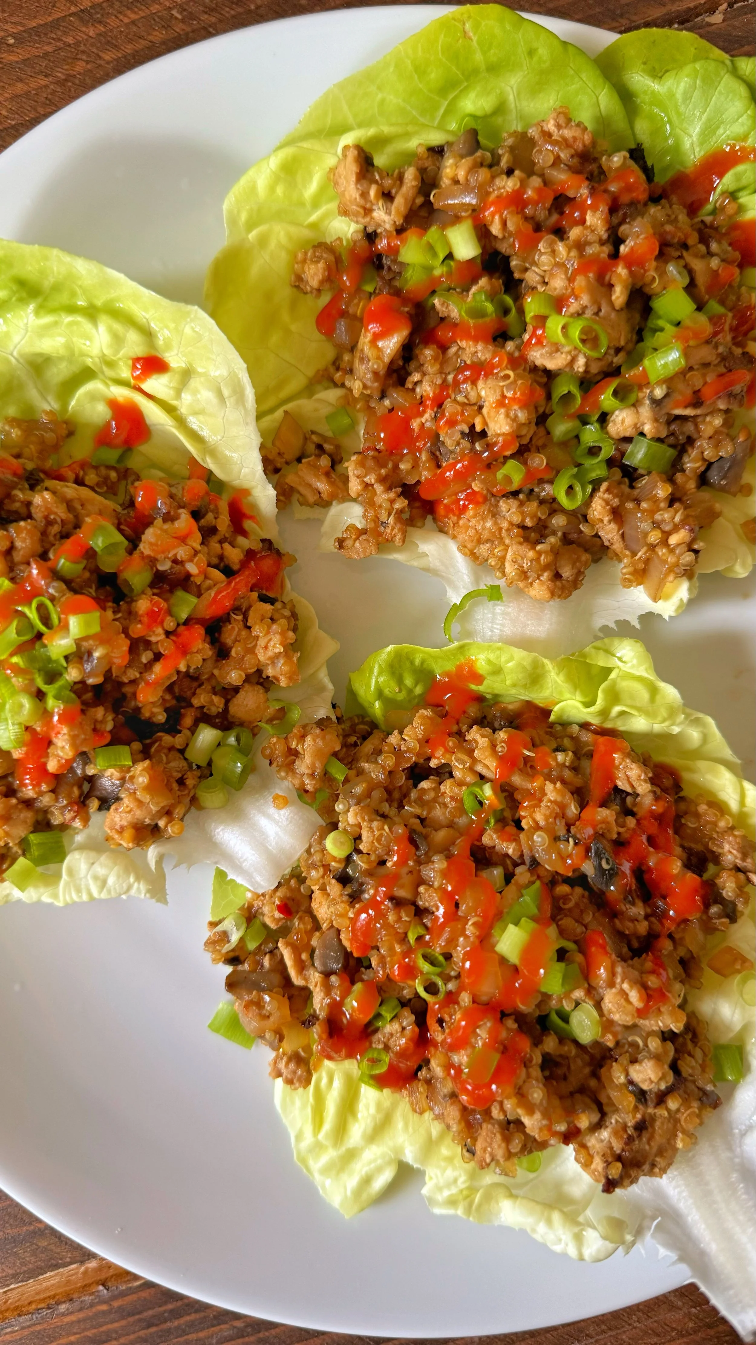 A close up shot of a plate of ground chicken on a bed of lettuce topped with chopped green onions and sriracha sauce.
