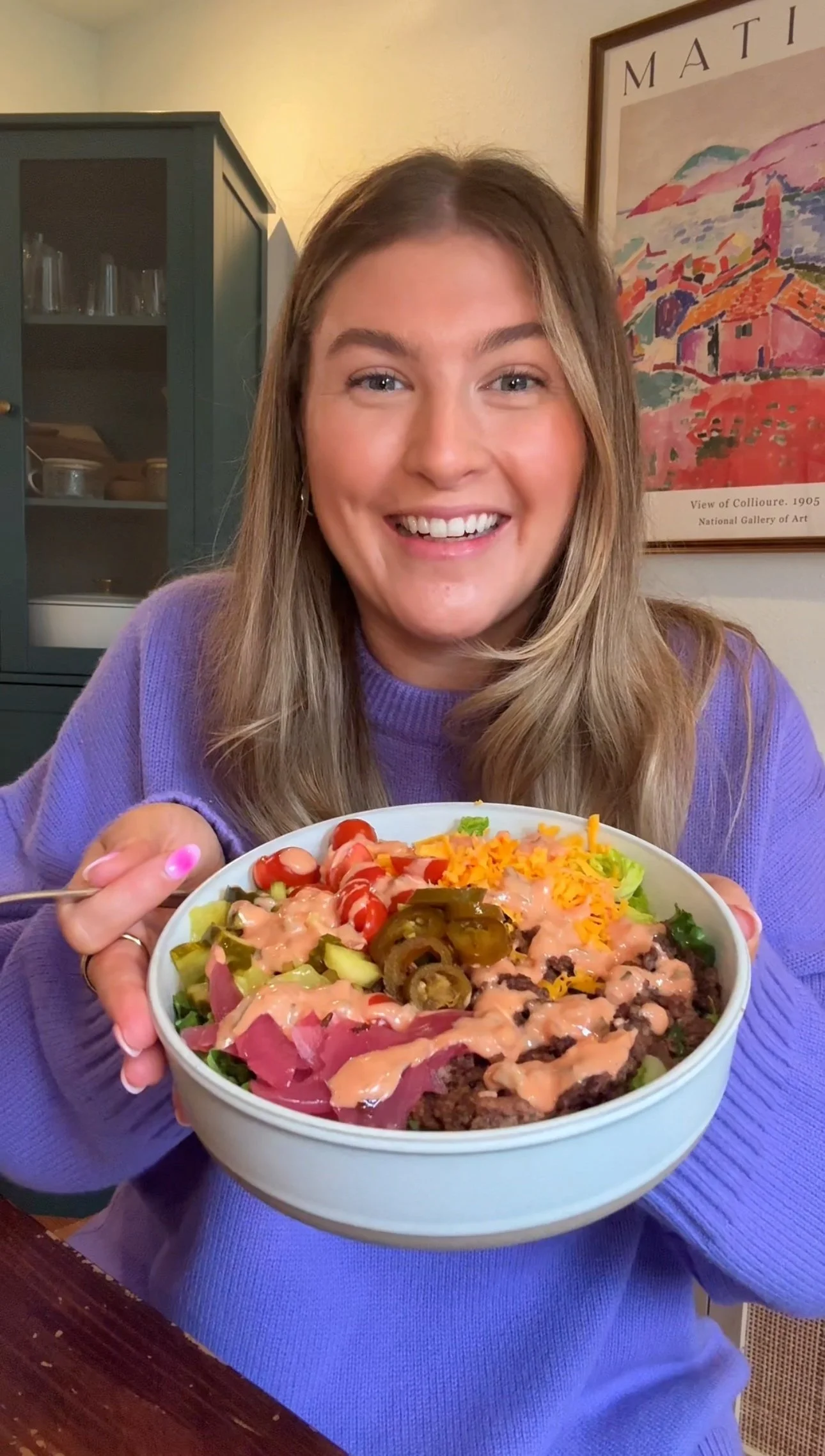 Cailee holding a  bowl of ground beef, pickled onions, tomatoes, grated cheese on a bed of lettuce topped with jalapeños and salad dressing. 