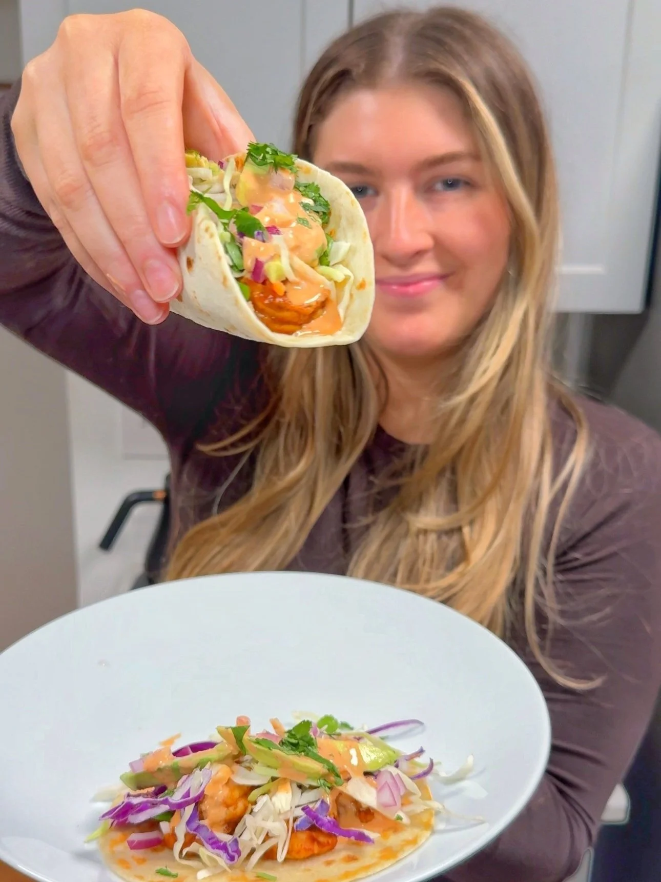 A shrimp taco with cabbage slaw on a plate with dressing on top. Another taco being held up in the air.