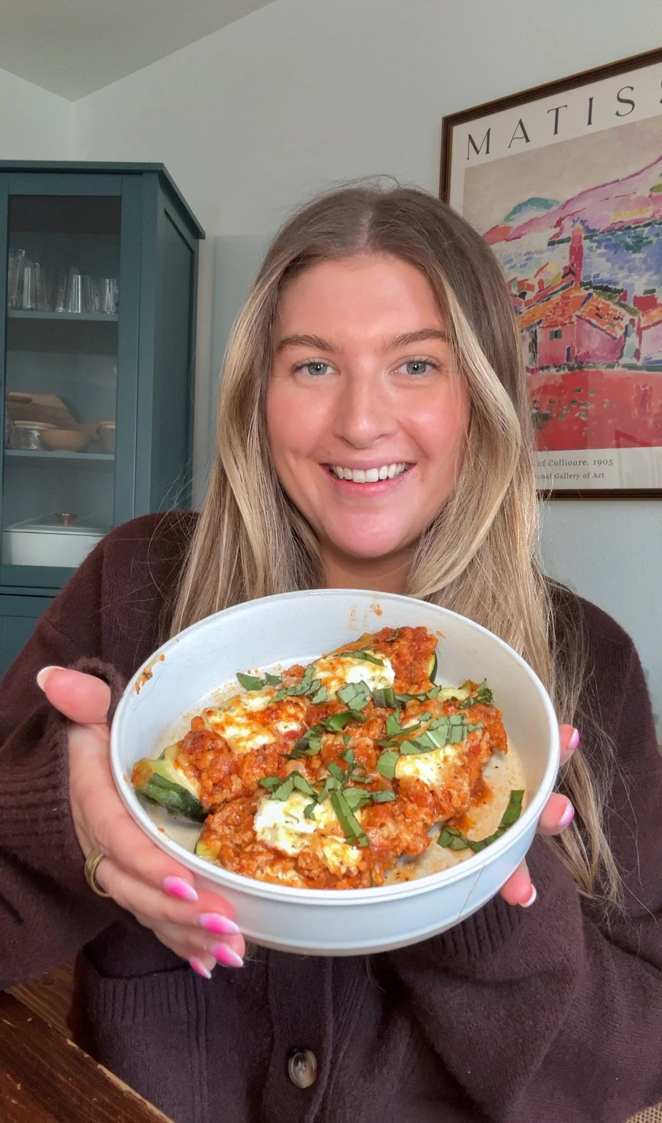 Cailee holding a bowl of stuffed zucchini with ground meat in a marinara sauce with cheese and fresh basil on top. 