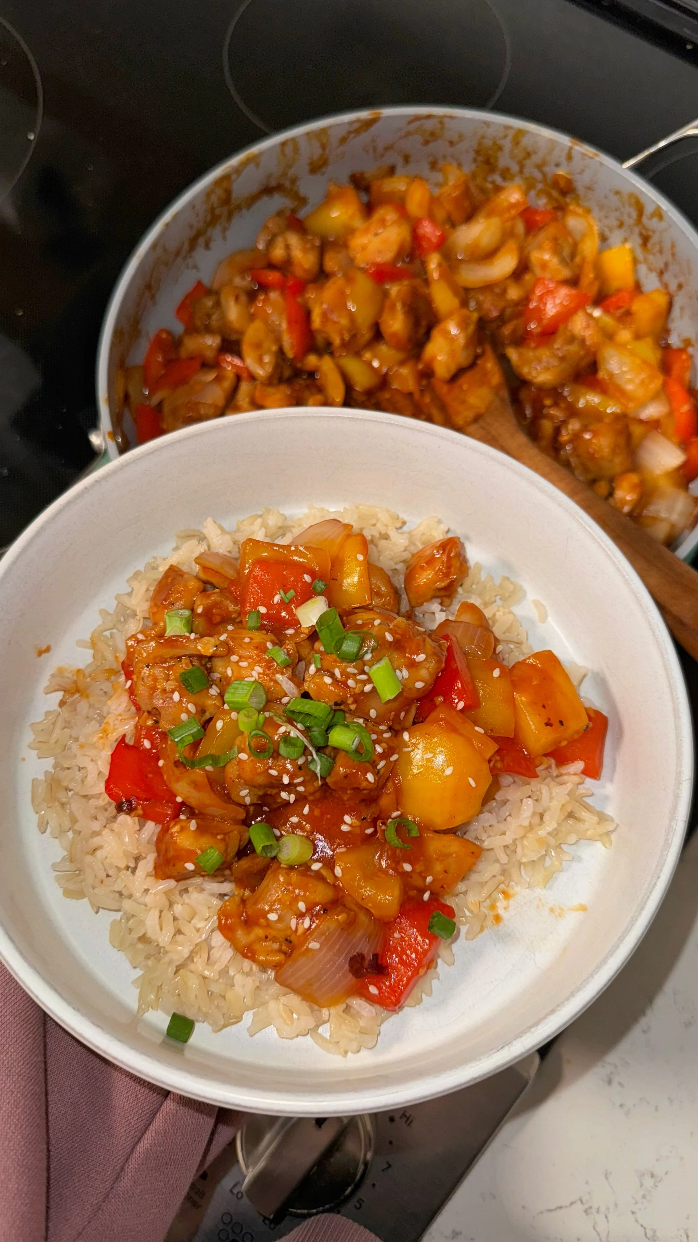 A bowl of sweet and sour chicken with pineapple chunks on a bed of rice topped with chopped green onions and sesame seeds.