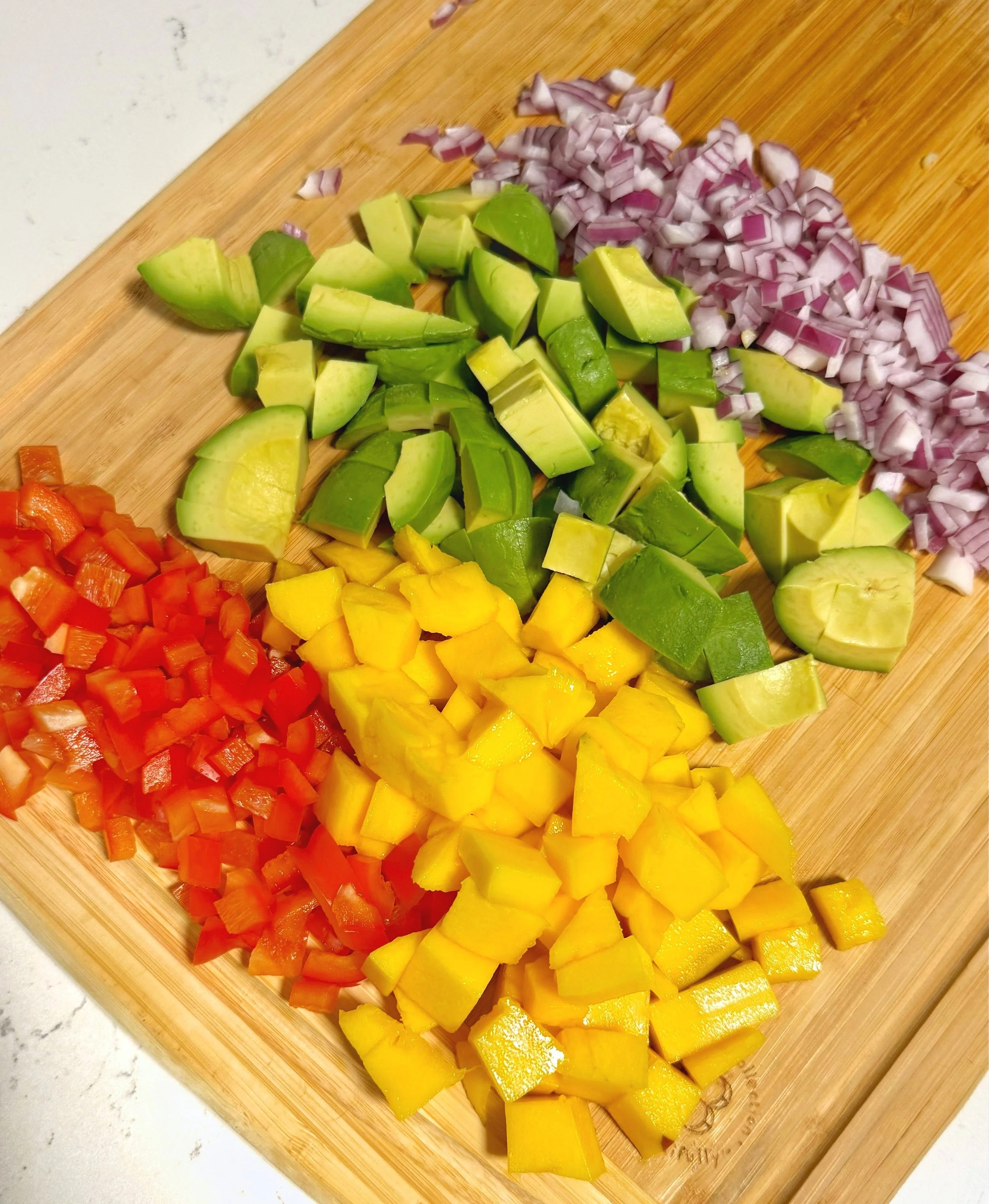 Chopped red peppers, red onions with chunks of avocado & mango on top of a cutting board. 