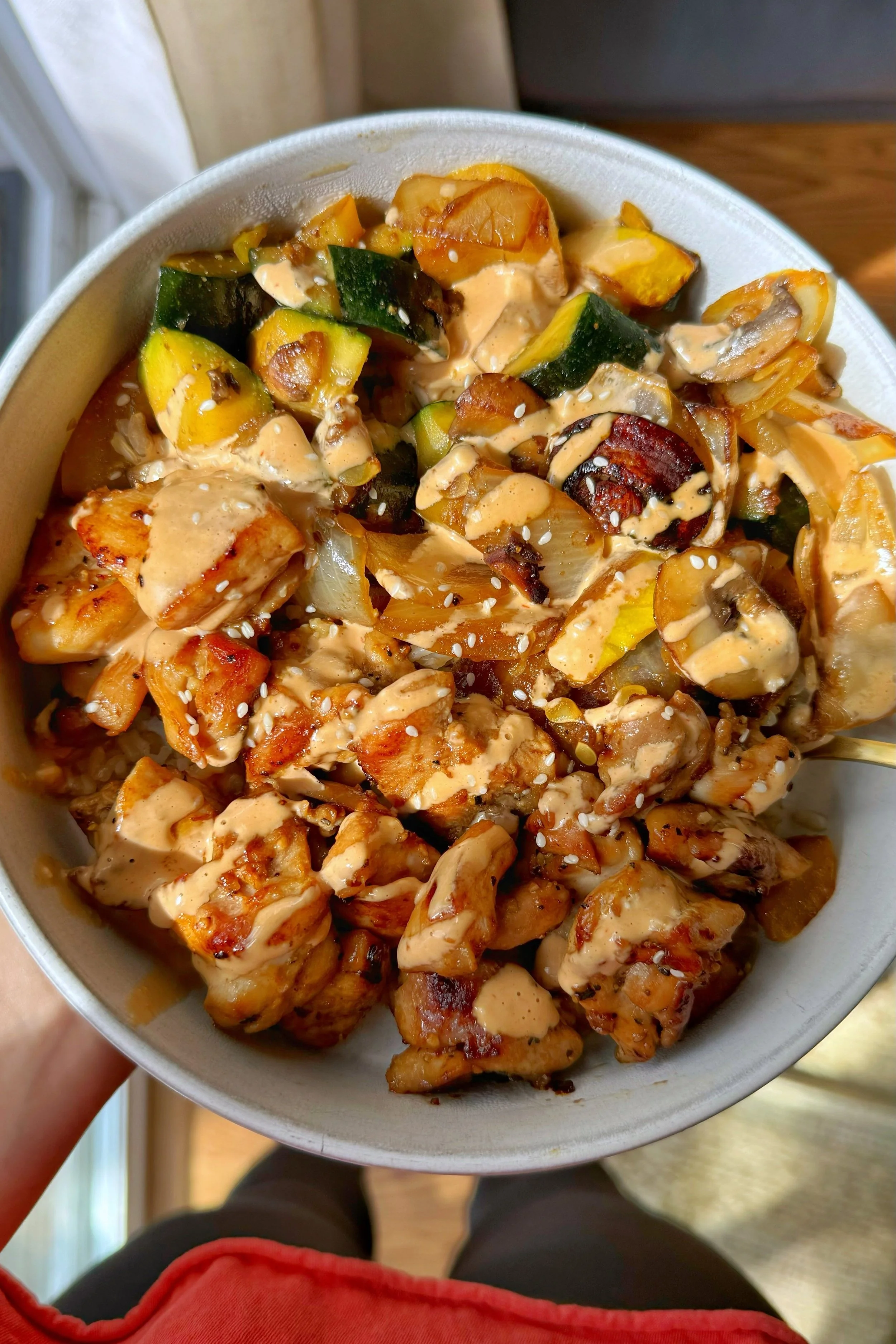 A bowl of chicken cubes with zucchini, summer squash, onions and sliced mushrooms served over rice with a drizzle of dressing and sesame seeds on top.