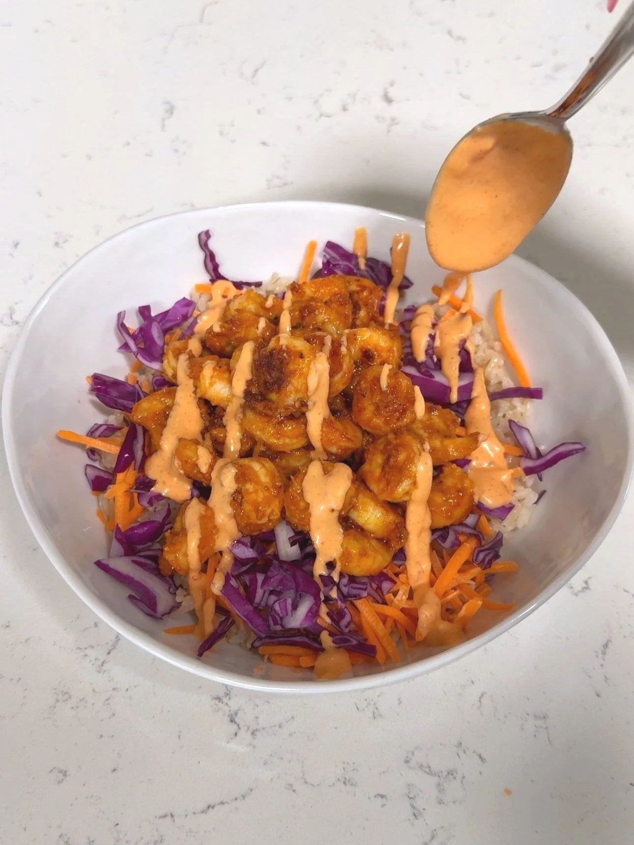 A bowl of spicy chicken over a bed of chopped purple cabbage, shredded carrots over brown rice topped with a dressing and chopped green onions. 