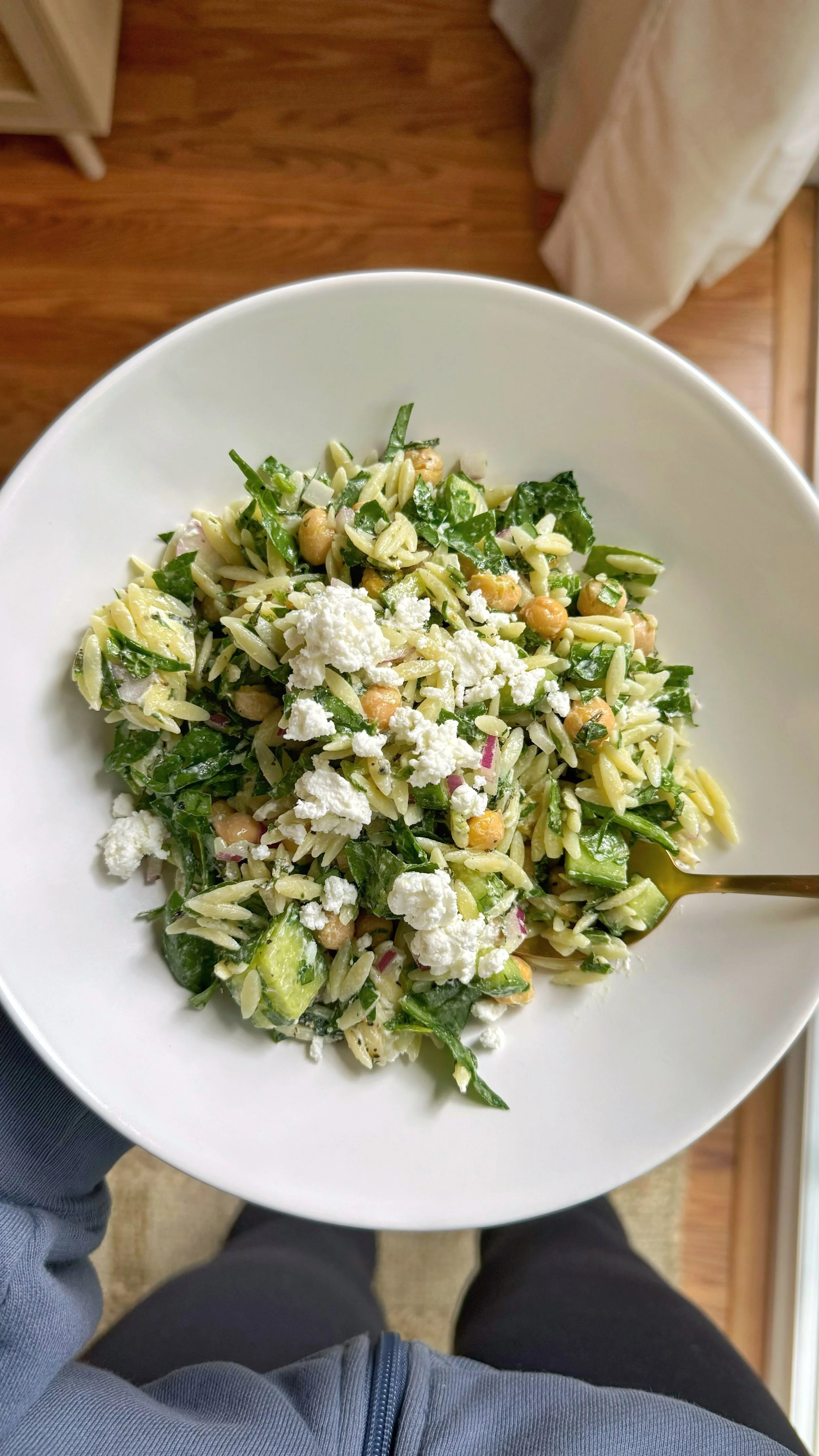A bowl of orzo salad with garbanzo beans, diced cucumbers, baby spinach, mint, and crumbled goat cheese topped with cracked pepper. 