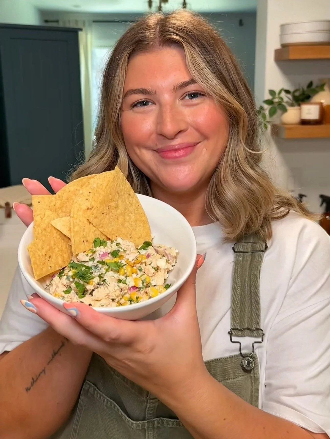 A bowl being held up with chopped chicken, corn, red onions, crumbled Cotija cheese topped with fresh cilantro. A side of chips are in the bowl.