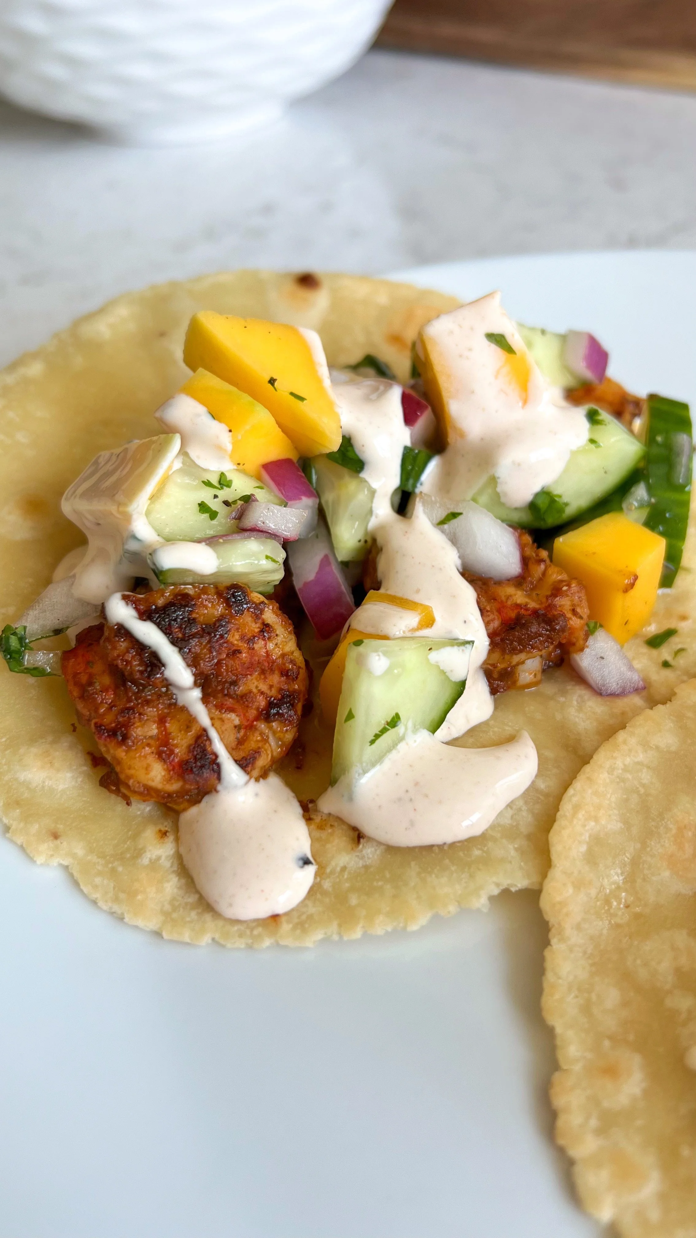 A close up shot of a taco shell with shrimp, mango, cucumbers, red onion with cilantro and a creamy lime sauce on top.