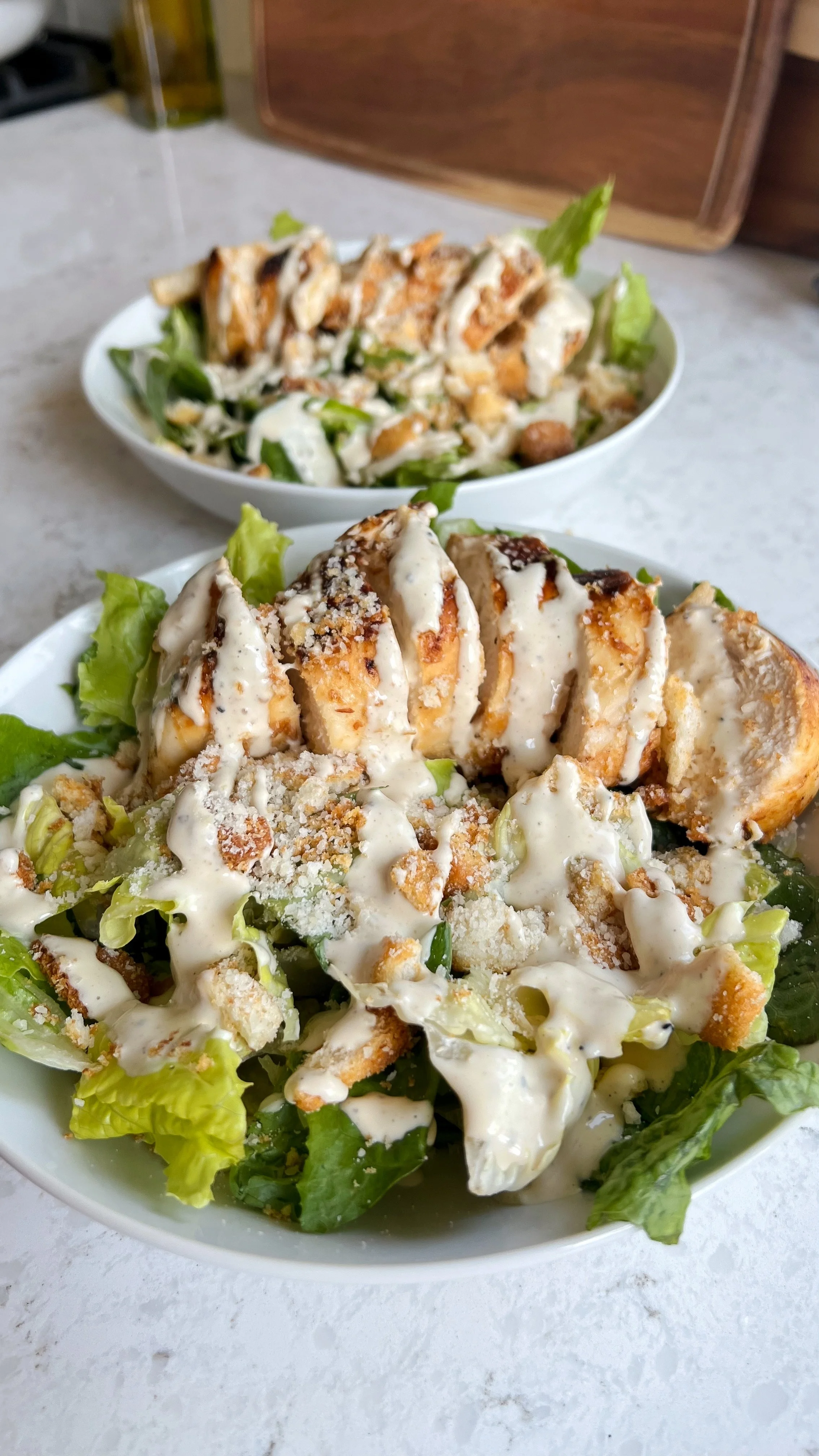 Two bowls of romaine lettuce, chicken, croutons, parmesan cheese with dressing on top.