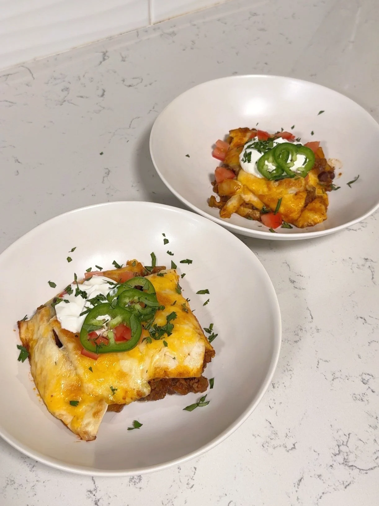 Two bowls each having an enchilada in them with sour cream, cilantro and fresh jalapeños.