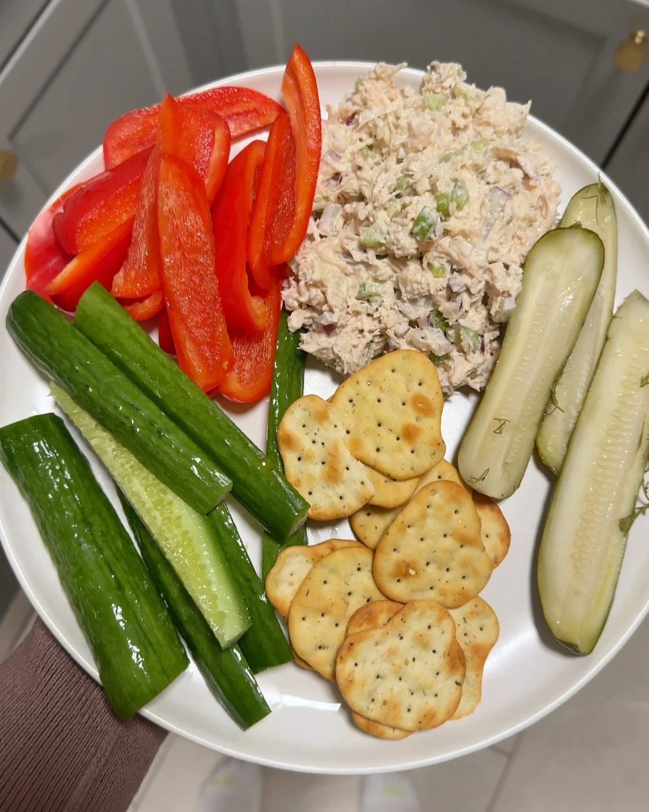A plate of chicken salad served with red peppers, cucumbers, pickles and crackers.