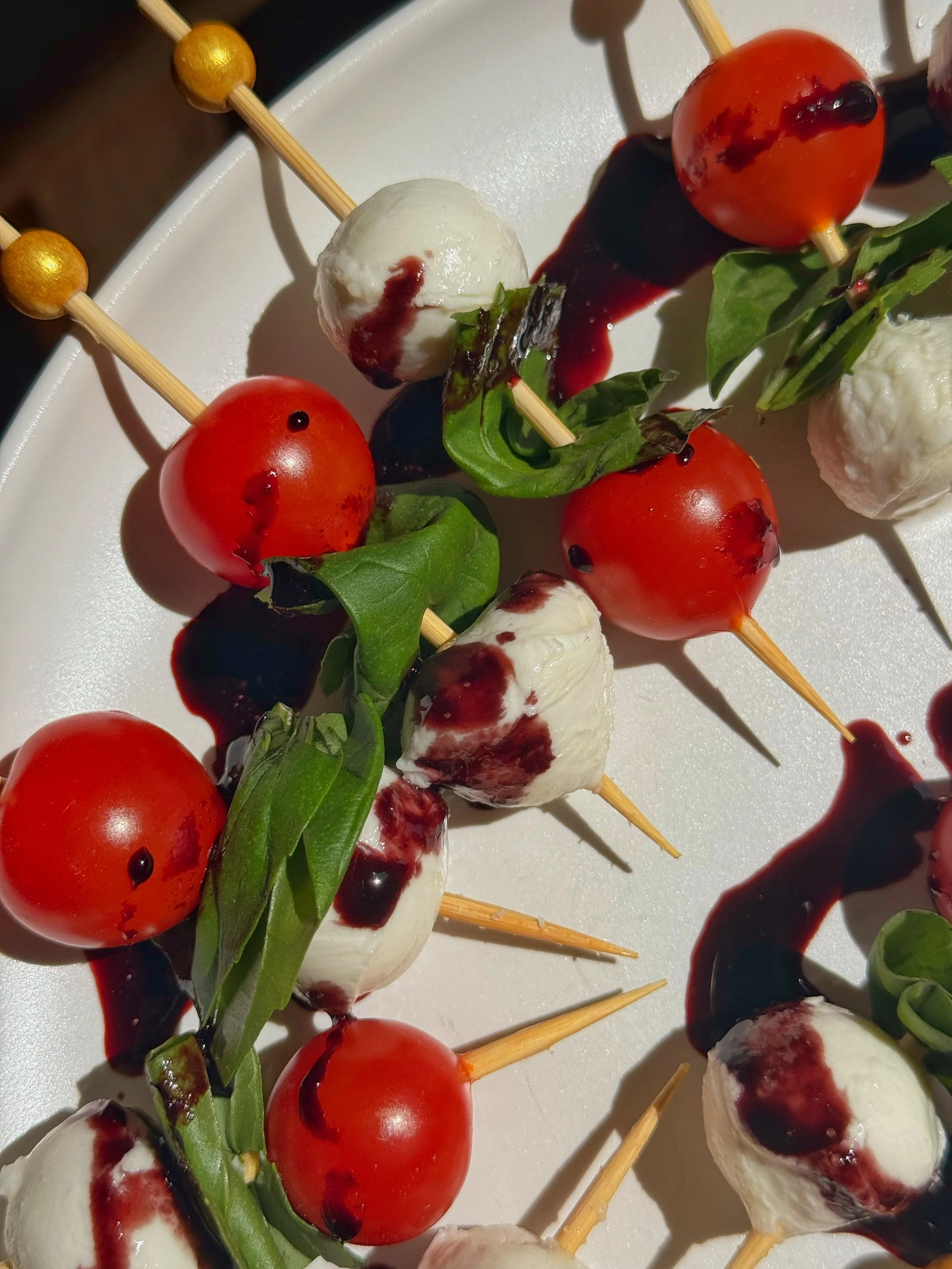 A close up shot of a plate with toothpicks that have tomato, basil, and mozzarella balls with a drizzle of blueberry glaze on top.