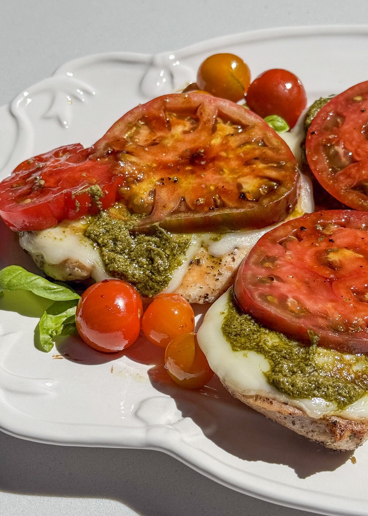 Pesto chicken topped with melted mozzarella and sliced heirloom tomatoes on a white plate.